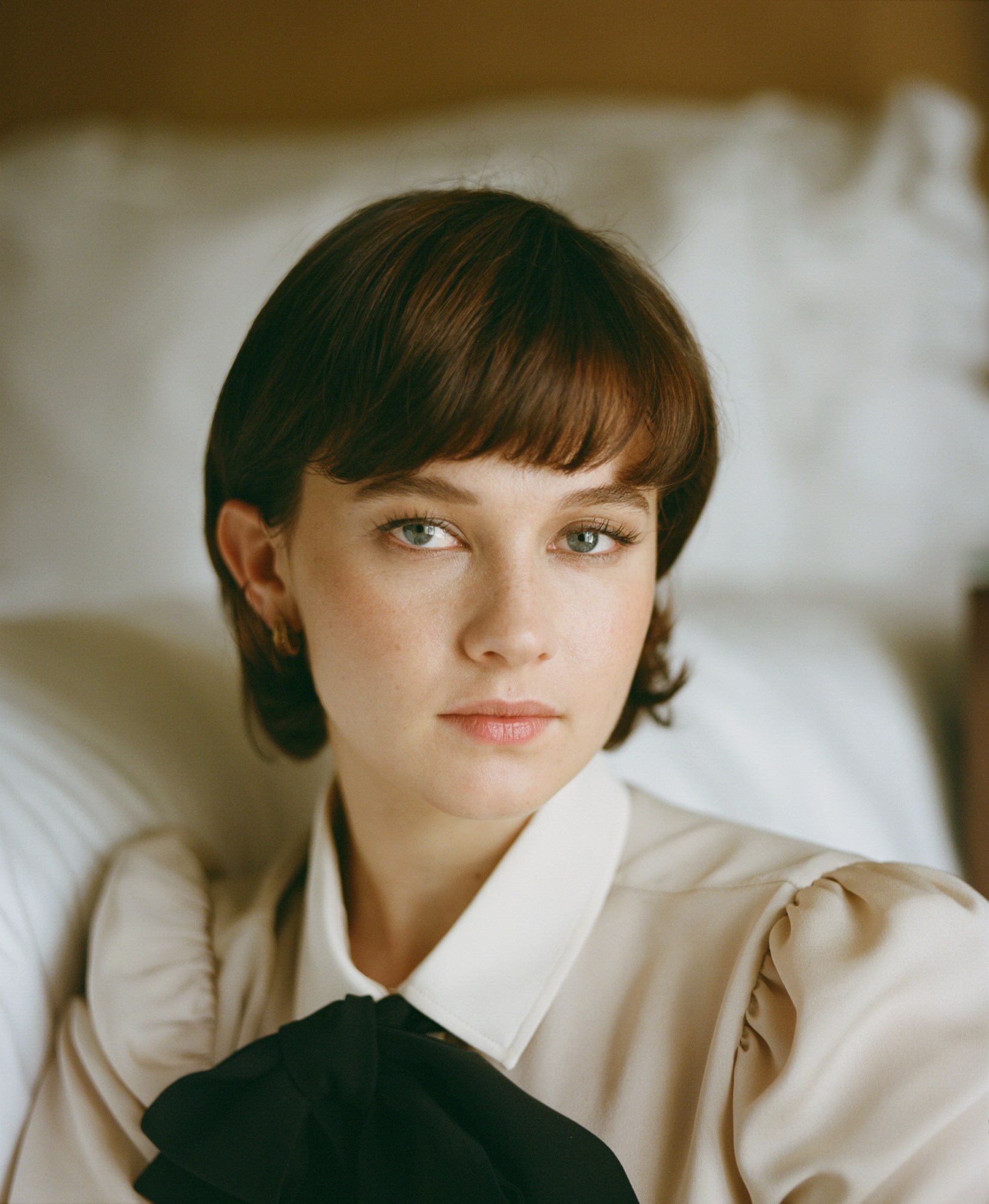 actress cailee spaeny wearing a cream shirt with a bow in the collar, leaning on a bed