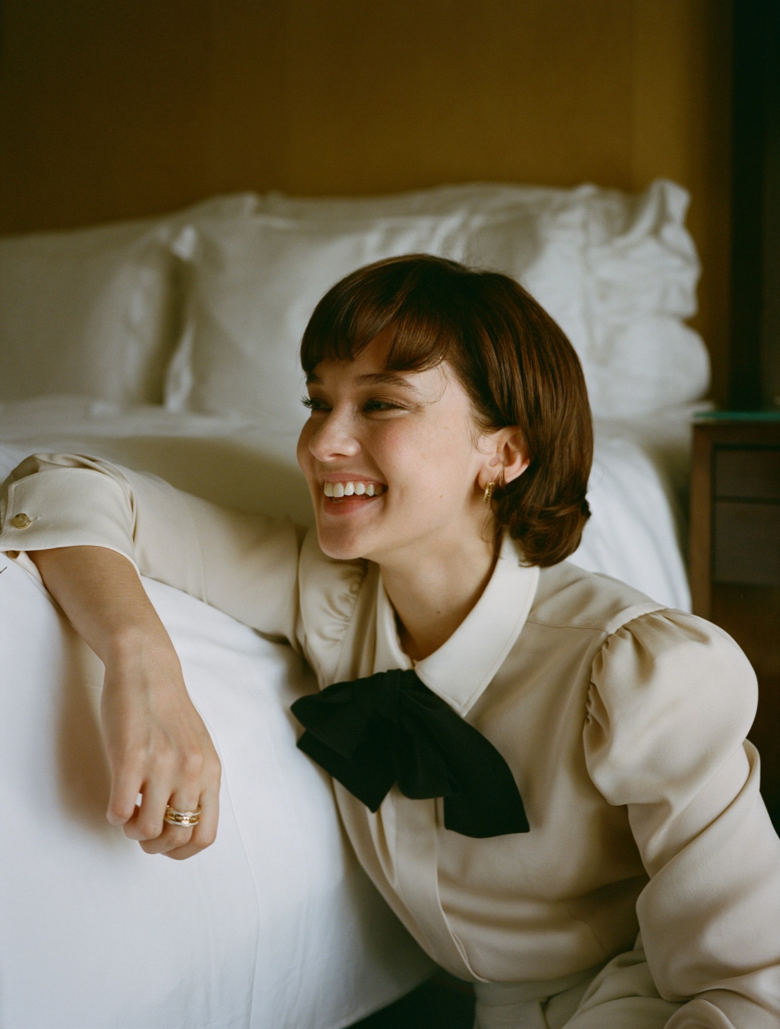 actress cailee spaeny wearing a cream shirt with a bow in the collar, leaning on a bed and smiling off to the side