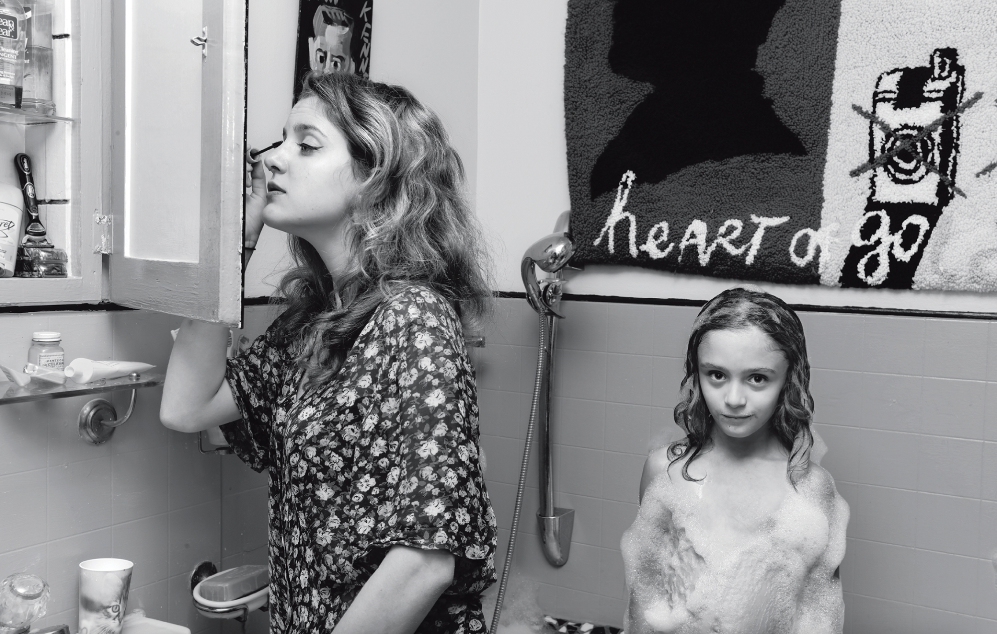 Photograph by Jim Goldberg of a woman doing her makeup in the bathroom cabinet mirror while a younger girl takes a bath from 'Coming and Going'