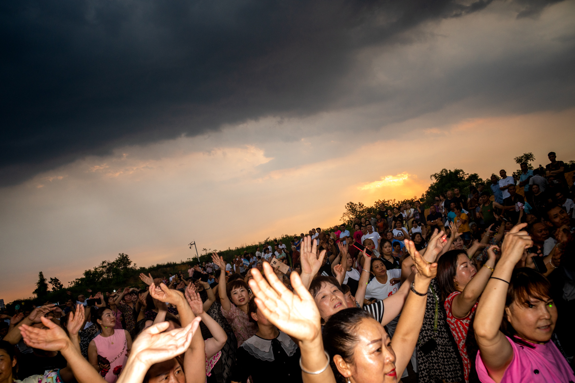 a shot of hundreds of older chinese people at an outside rave, arms in the air as the sun sets