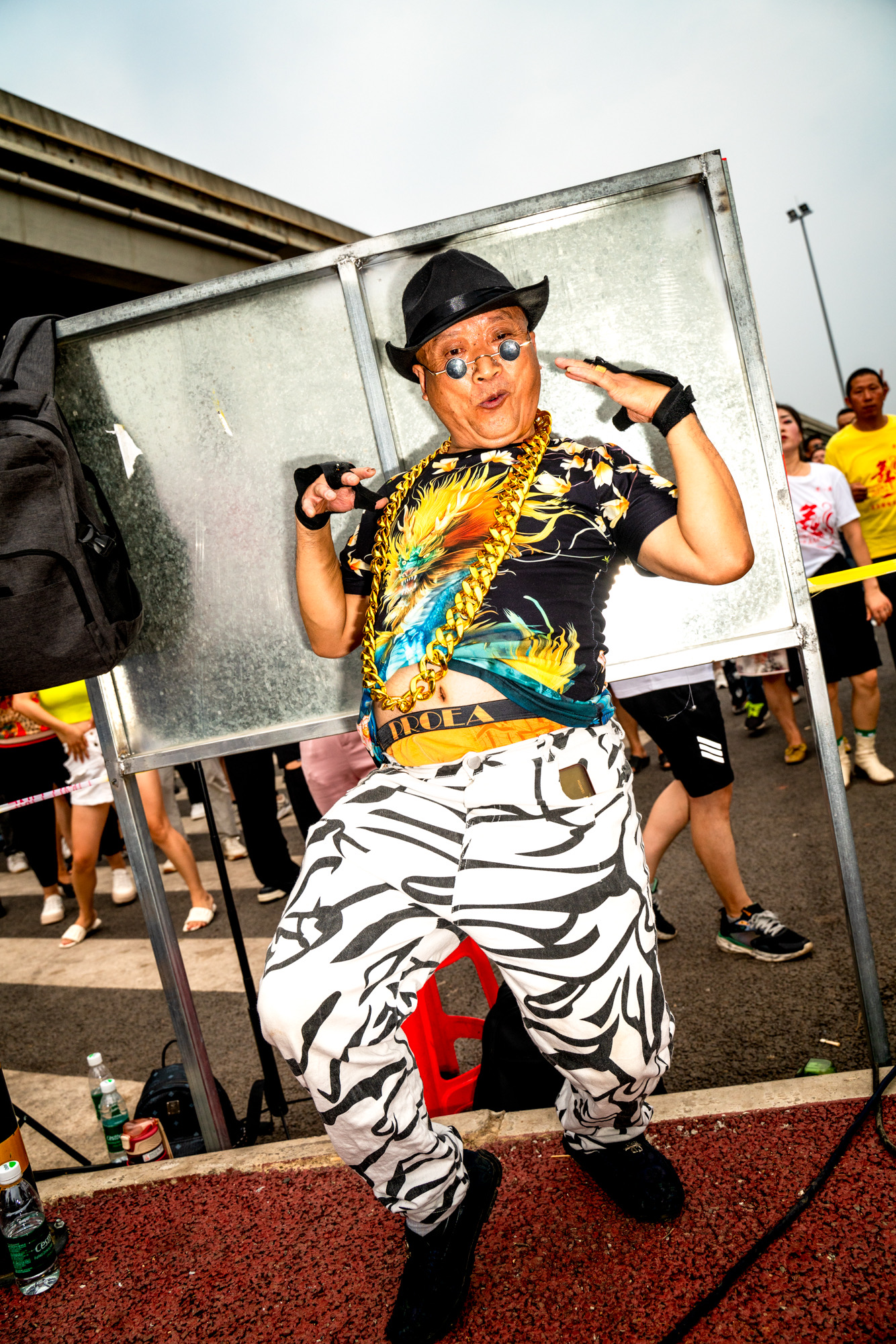 an older chinese man in zebra print jeans, an oversized gold chain and tiny round sunglasses performs for the camera