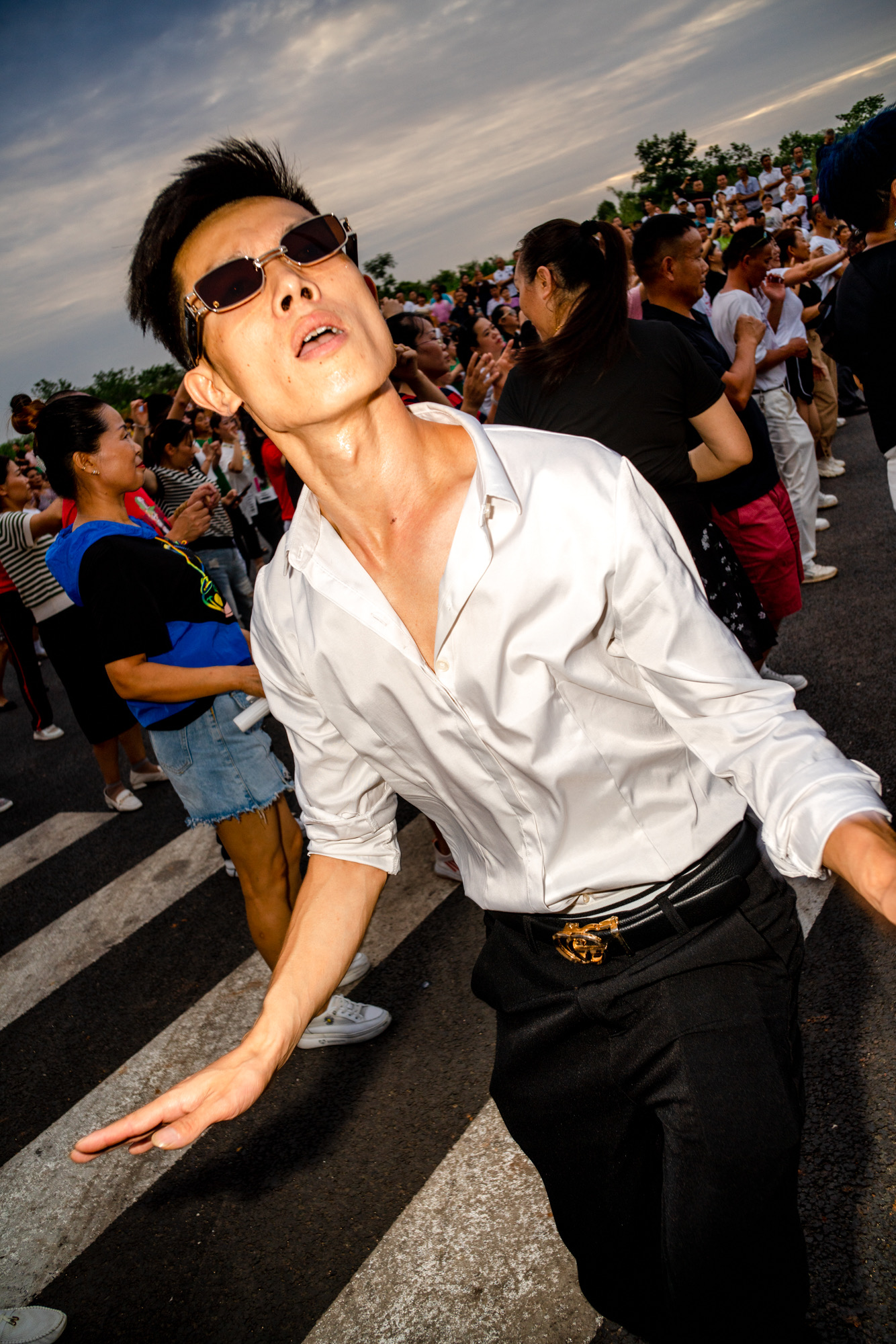 a man in a white silk shirt, black jeans and angular sunglasses dances outside in a crowd at dusk