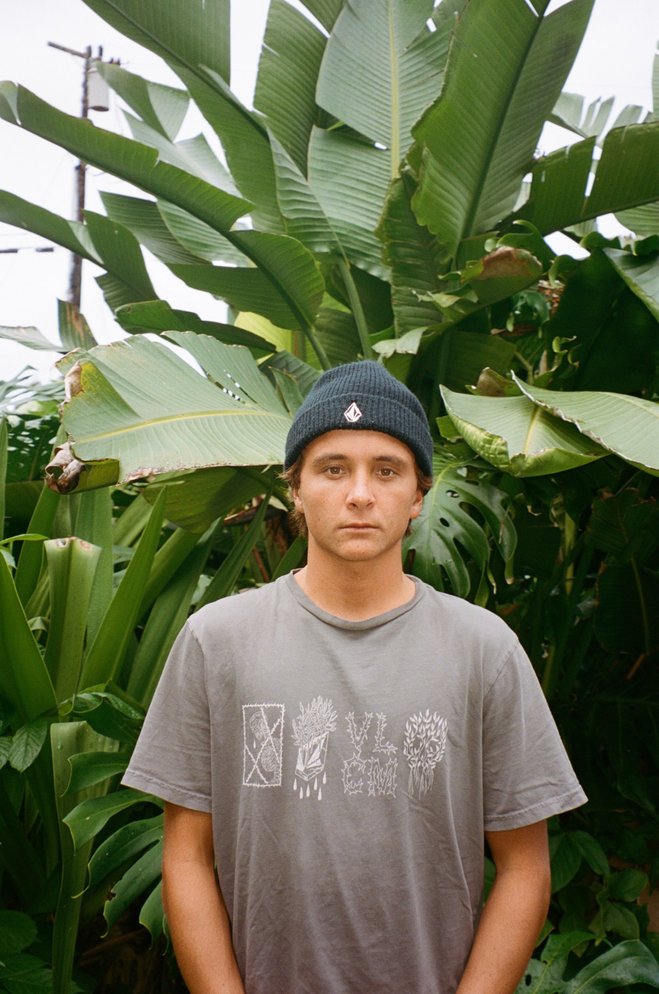 a young man in a faded surf tee and beanie stands in front of a wall of foliage