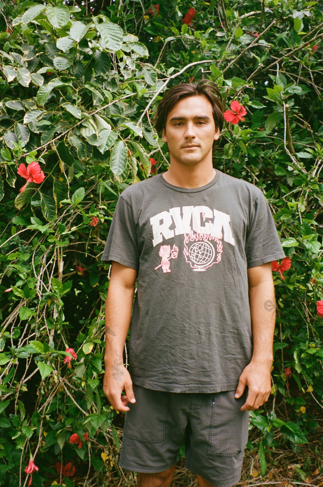 a tan young man in a faded grey tshirt and board shorts stands in front of a leafy bush peppered with bright pink flowers; he has floppy brunette hair and tattoos on his forearms