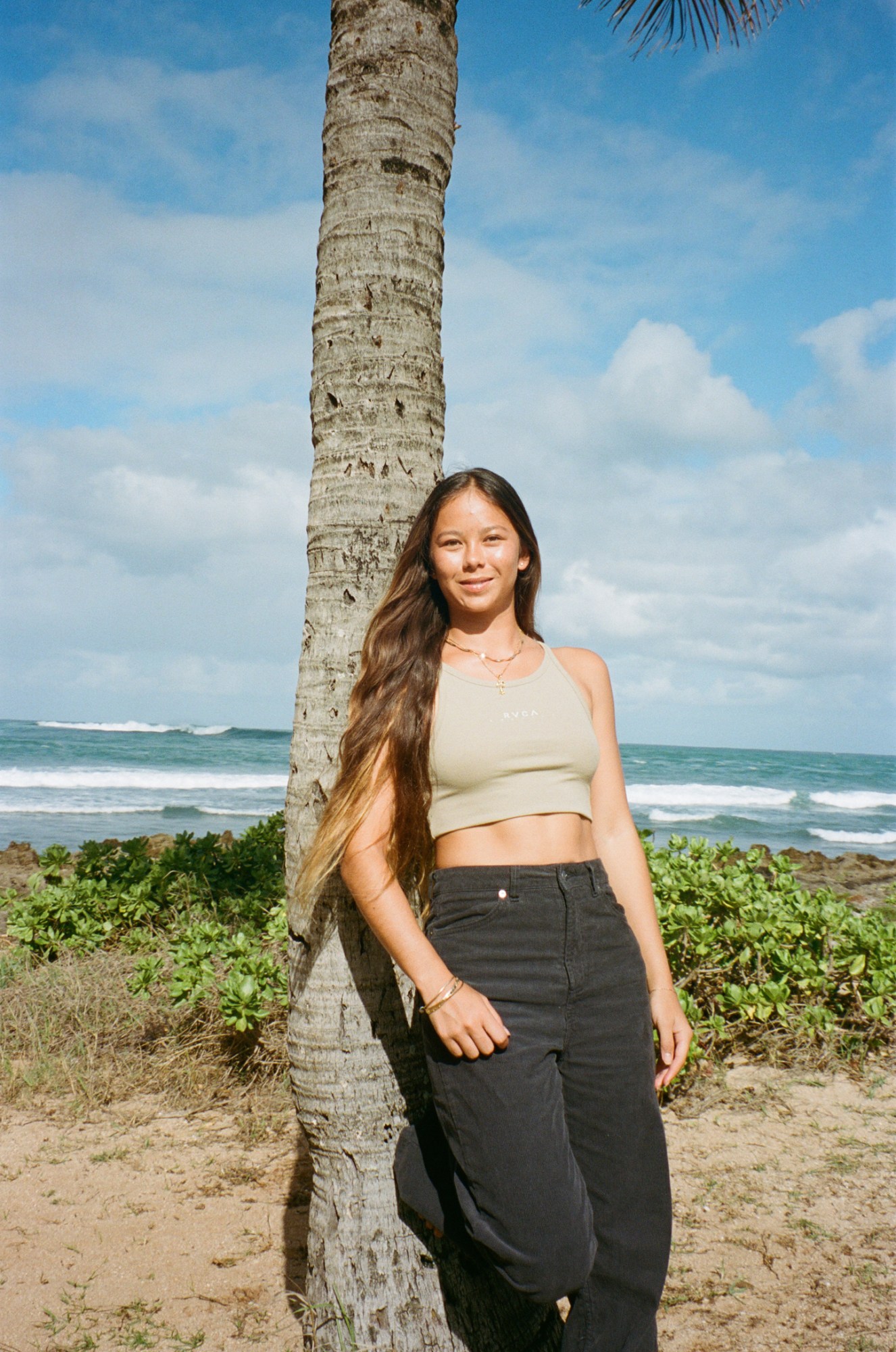 a young woman with very long brunette hair leans against a palm tree overlooking the sea; she's wearing dark jeans and a cropped tank top