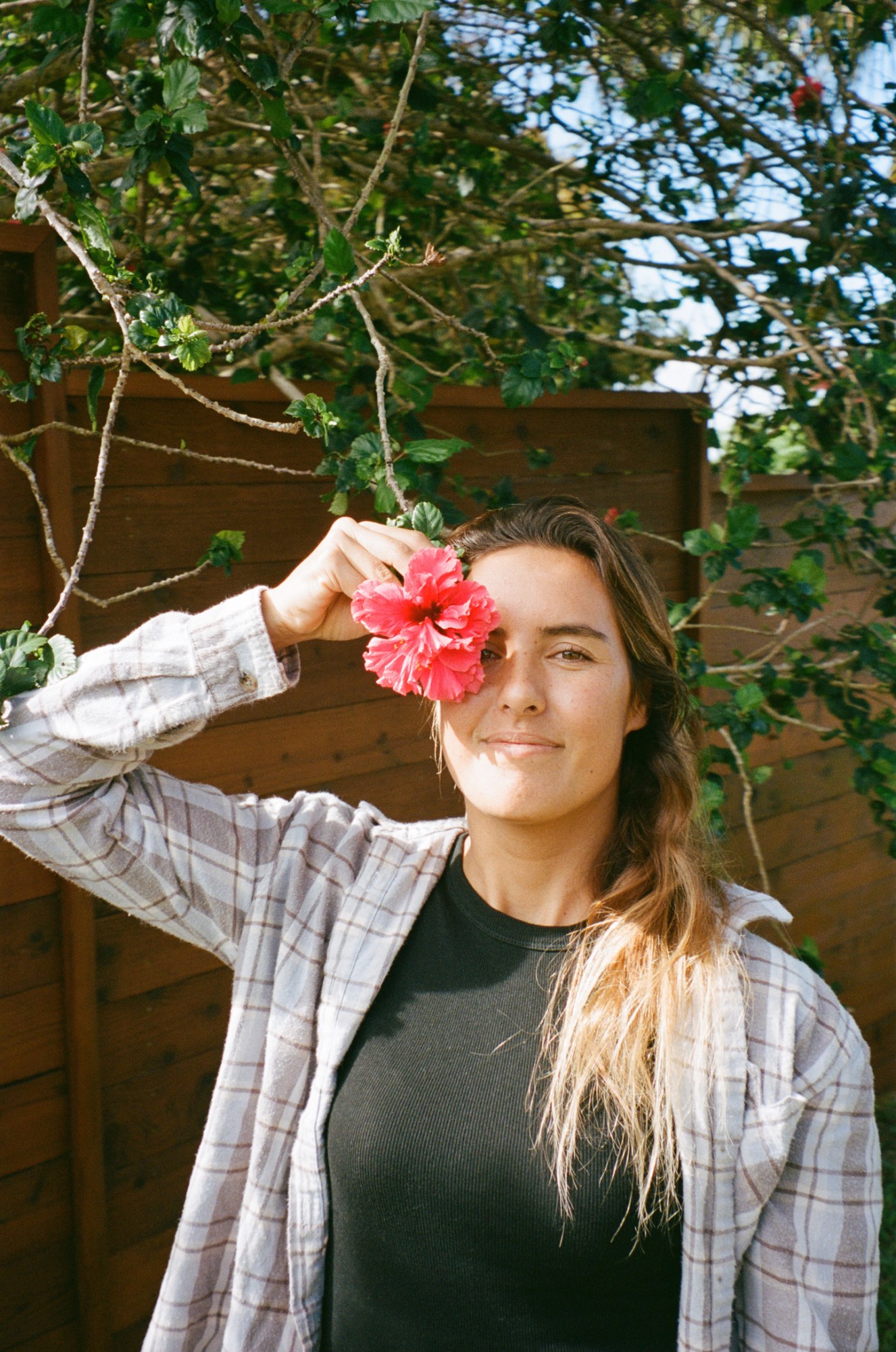 a young brunette woman in a checked shirt smiles as she holds a pink tropical flower over her right eye