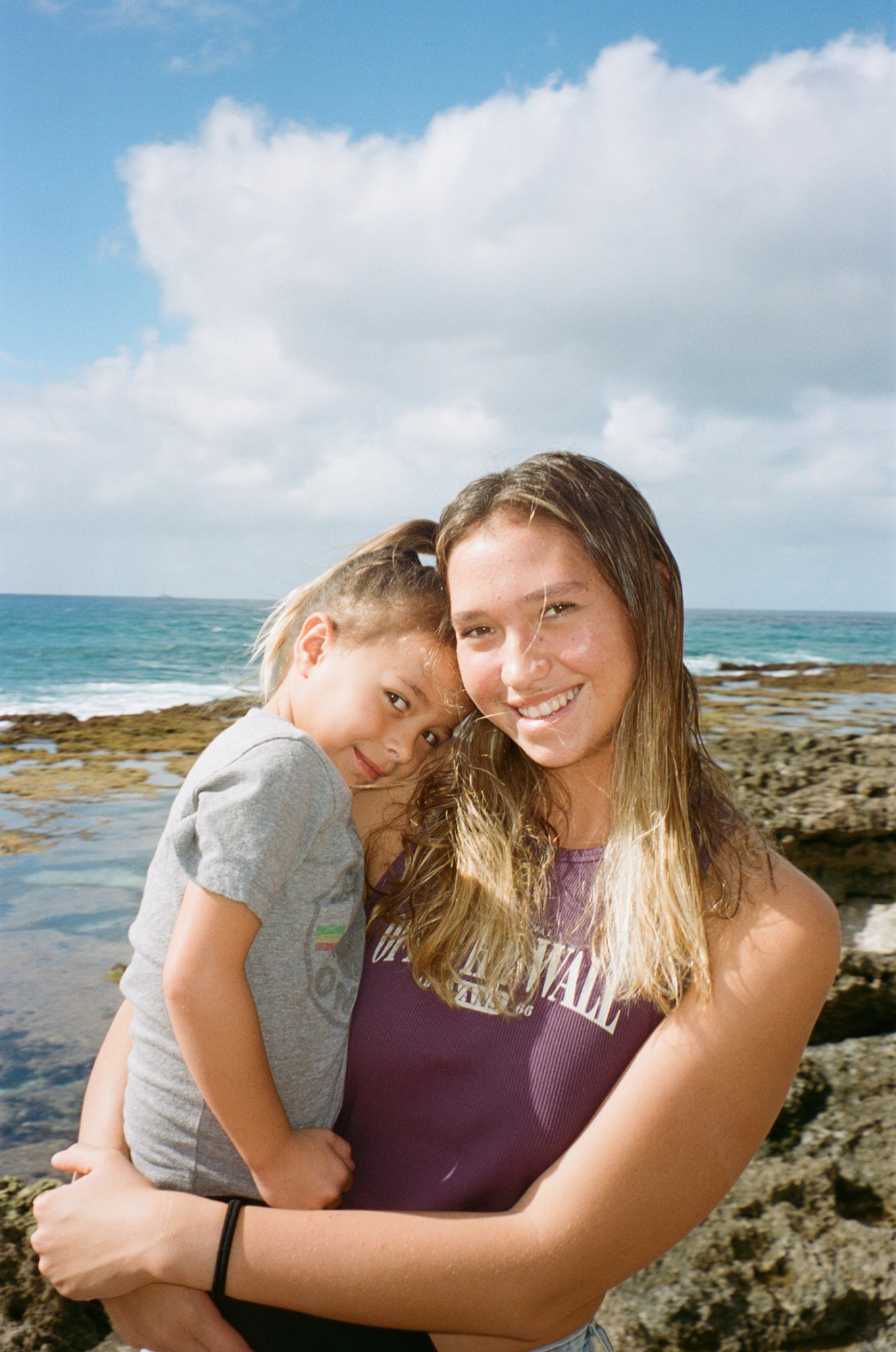 a young blonde surfer smiles on a rocky shore, holding who appears to be her younger sister in her arms