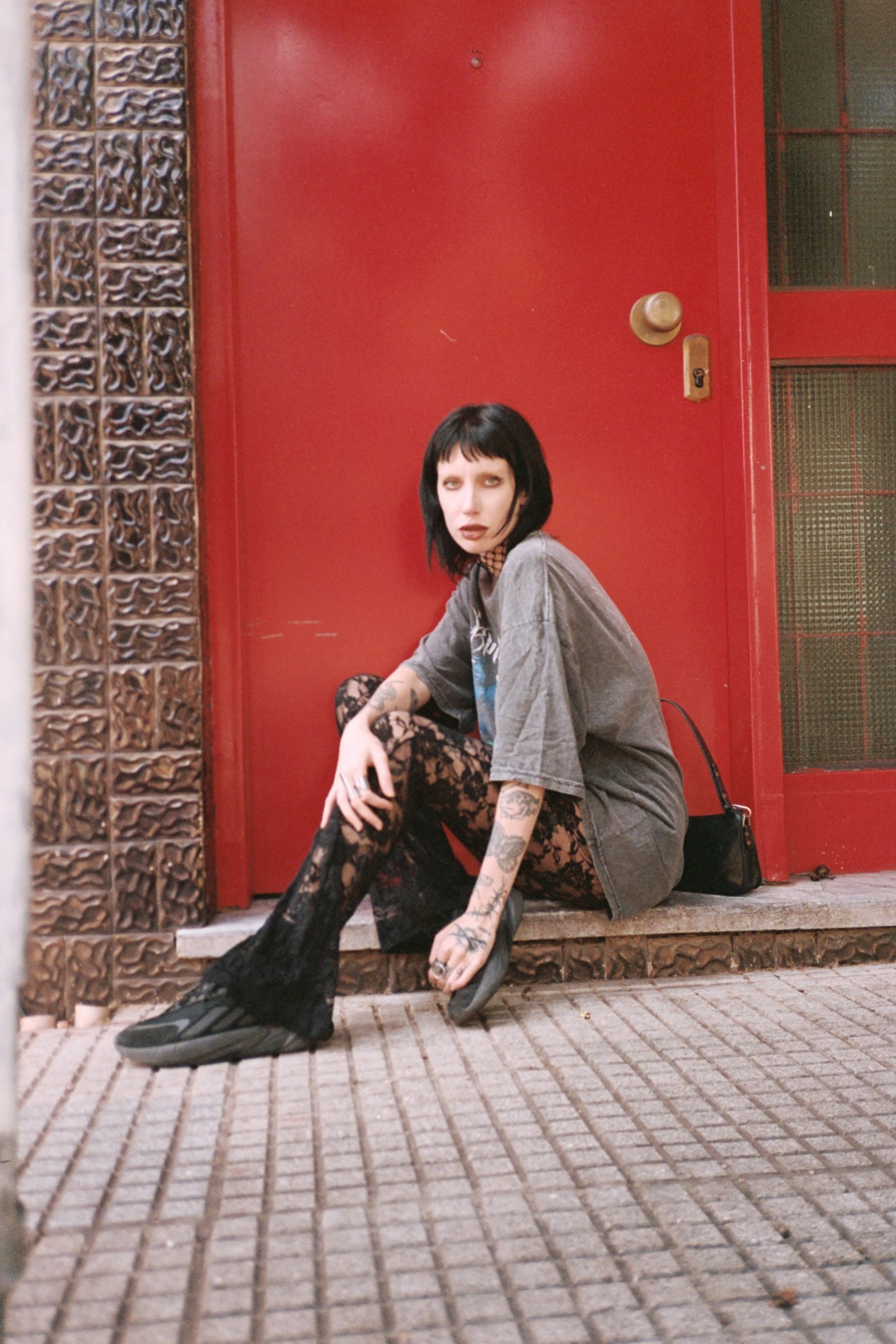 a gothy woman with black hair and a short fringe sits on a step in front of a bright red door