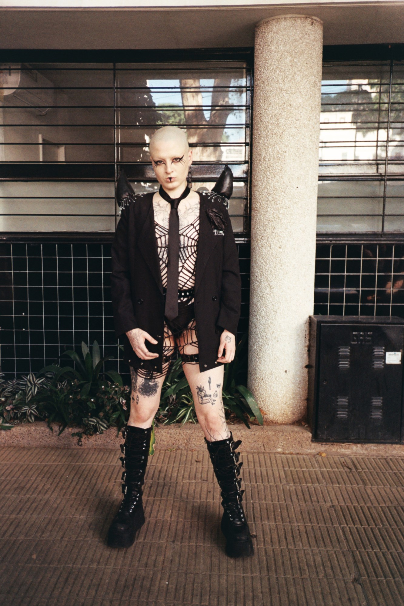 a goth wearing big boots, a spider web cut-out dress with visible underwear and a black blazer stands in the street