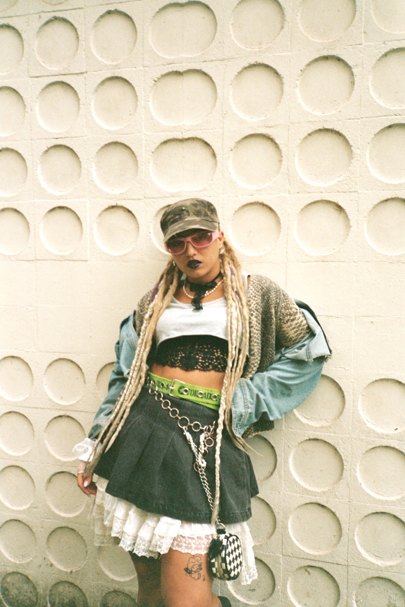 a woman in a frilly short kilt with long blonde dreads poses against a wall