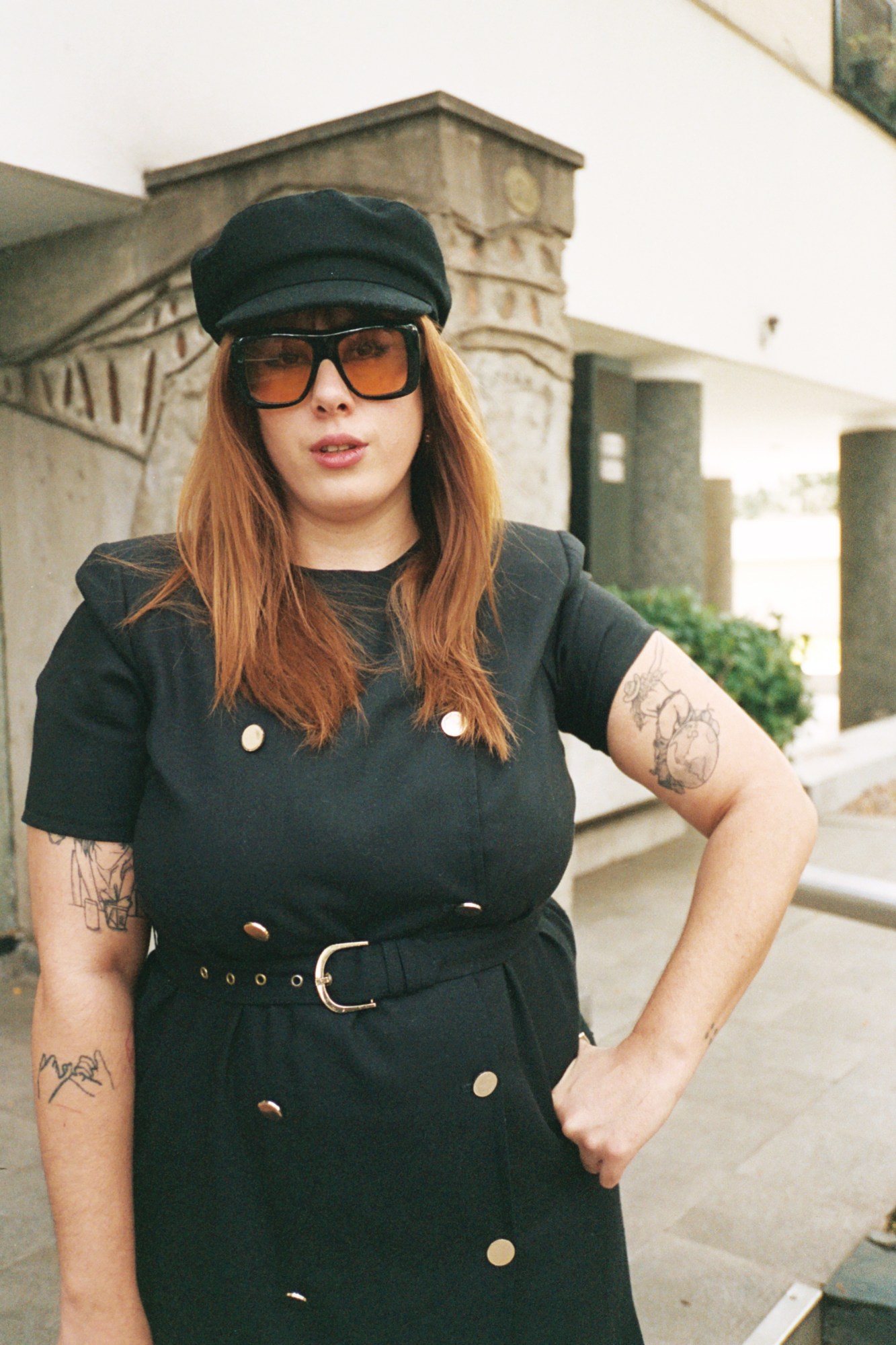 a woman in a black vintage dress, hat and orange-lensed sunglasses poses with her hand on one hip; her arms are covered in tattoos