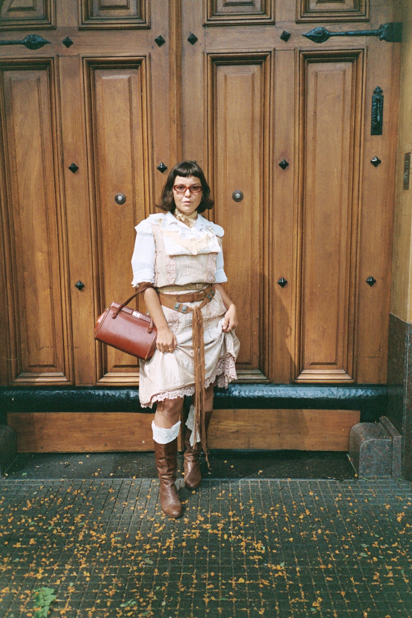 a woman in a pale pink lace dress with brown leather accessories stands in front of a large wooden door