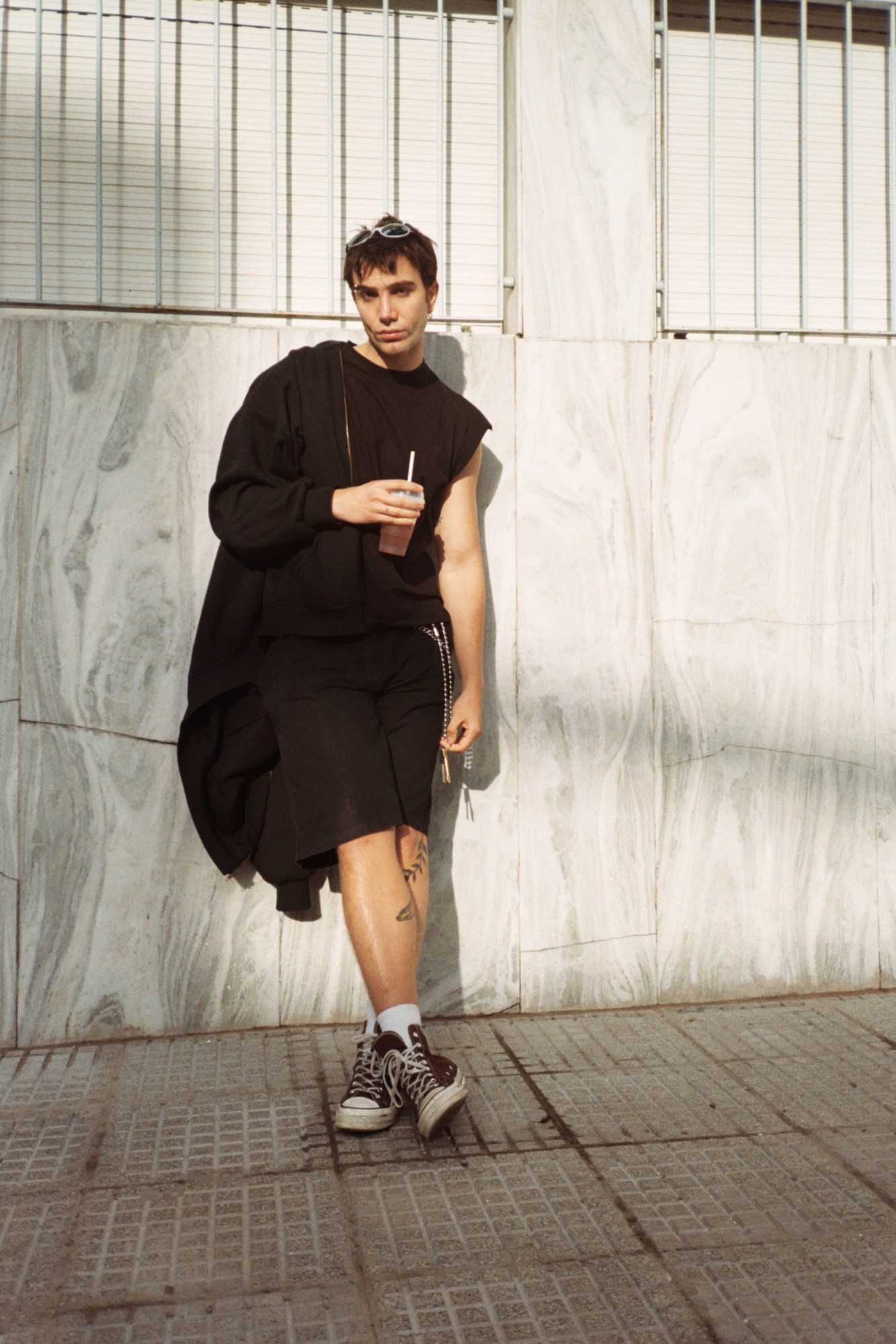 a person in black shorts and a tank top leans against a stone wall holding an iced drink