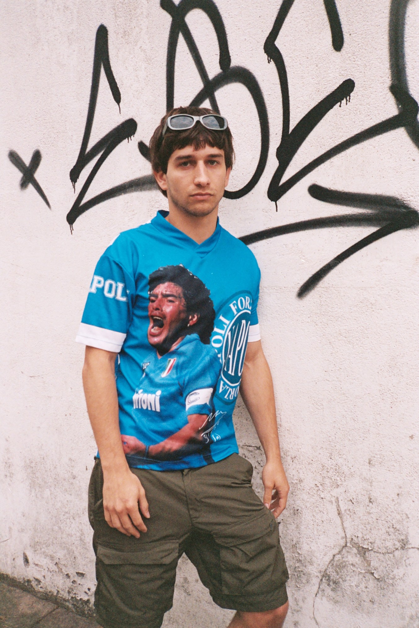 a man in a bright blue football top and combat shorts leans against a wall of graffiti