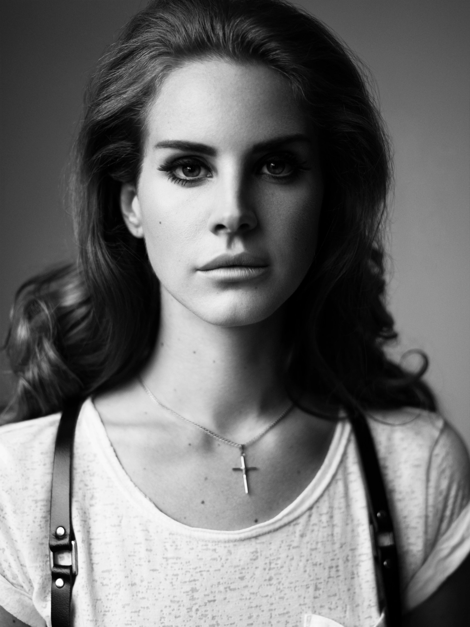 a headshot of young lana del rey in a white t-shirt, black harness and a cross necklace