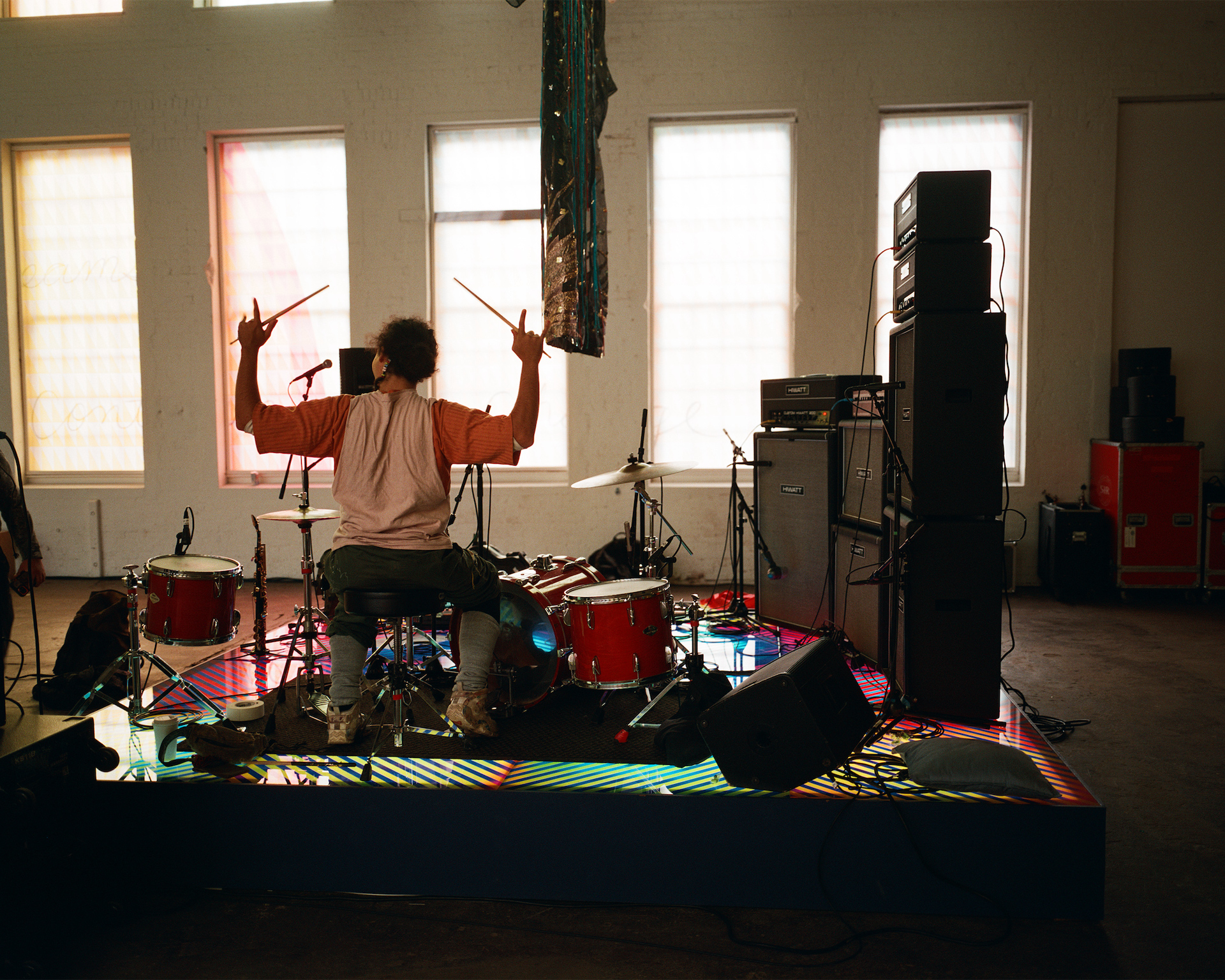 A musican is seen from behind, sitting at a drum kit with drum sticks in her hands.