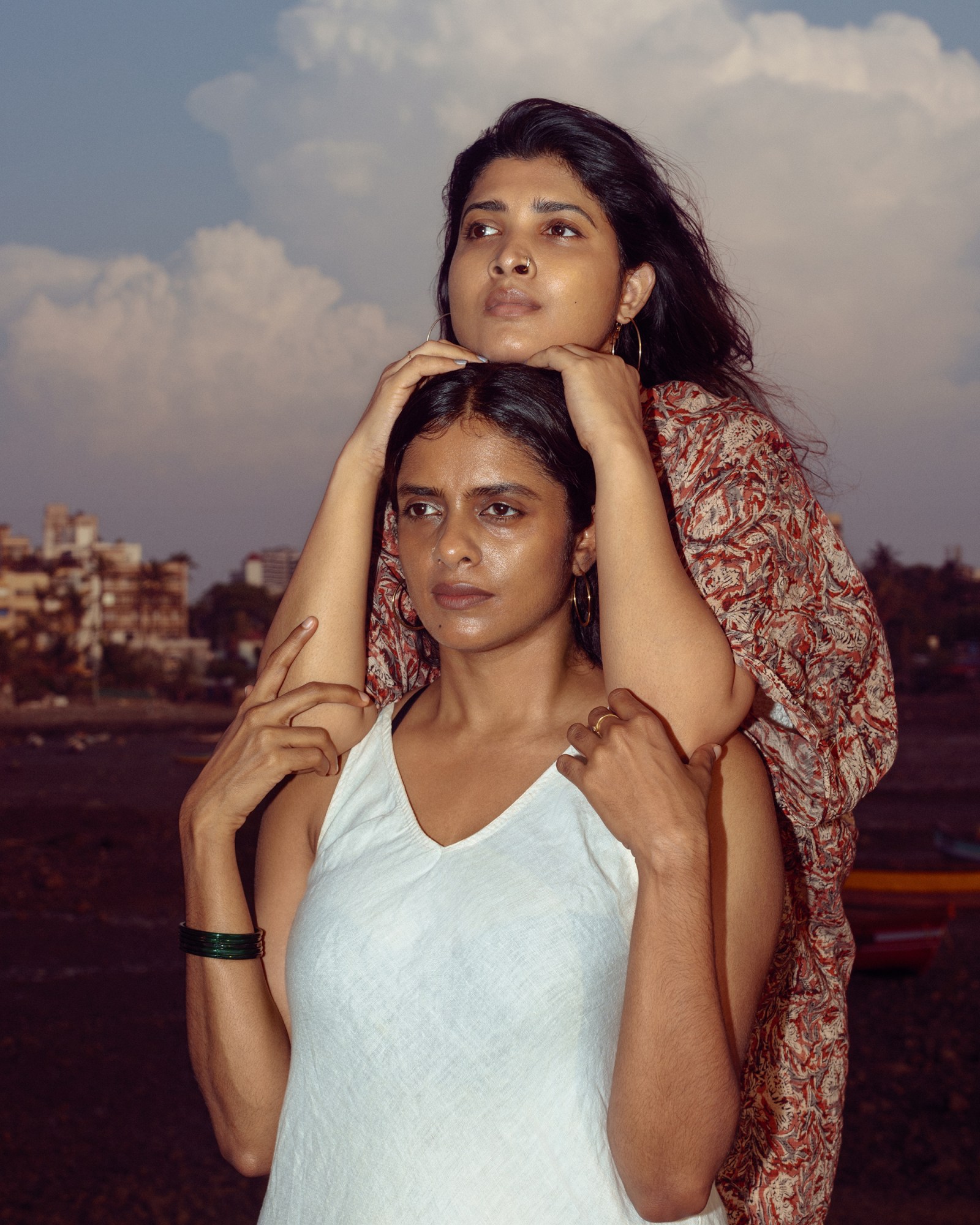 Indian All We Imagine as Light actors Kani Kusruti and Divya Prabha, both with dark brown hair, pose, looking off into the distance. Kusruti, a woman with long hair wearing a bracelet and a white slip dress, holds the shoulders of Prabha, with shoulder length dark hair wearing a patterned shawl, who rests her chin on Kusruti's head.