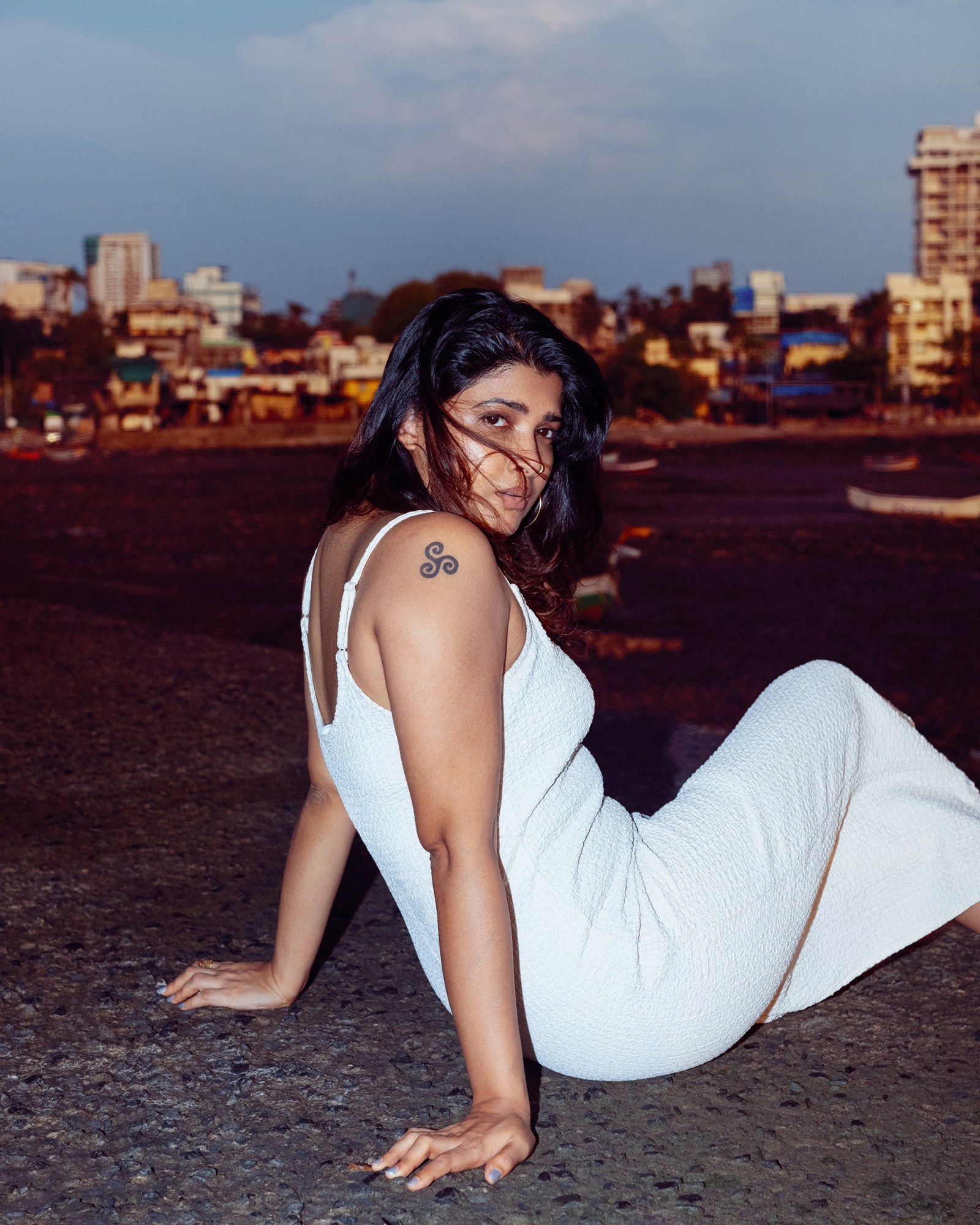 Indian All We Imagine as Light actor Divya Prabha, a brown Indian woman with shoulder length dark hair and a tattoo on her shoulder, sits on a shoreline looking over her shoulder to the camera.