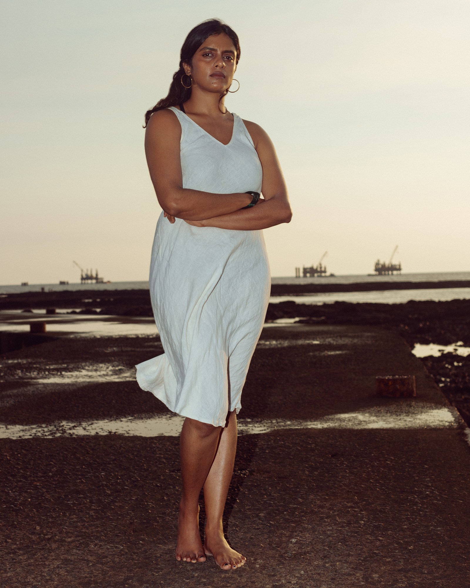 Indian All We Imagine as Light actor Kani Kusruti, a brown woman with long dark hair tied back stands in a white slip dress looking to the camera by the sea with her arms folded