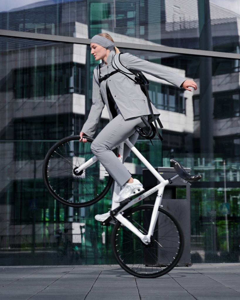 Woman does a wheelie on a bike wearing a grey suit.