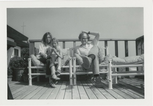 Joan Didion is pictured in a black and white photograph sitting on a deck, beside her husband John Dunne and their daughter Quintana.