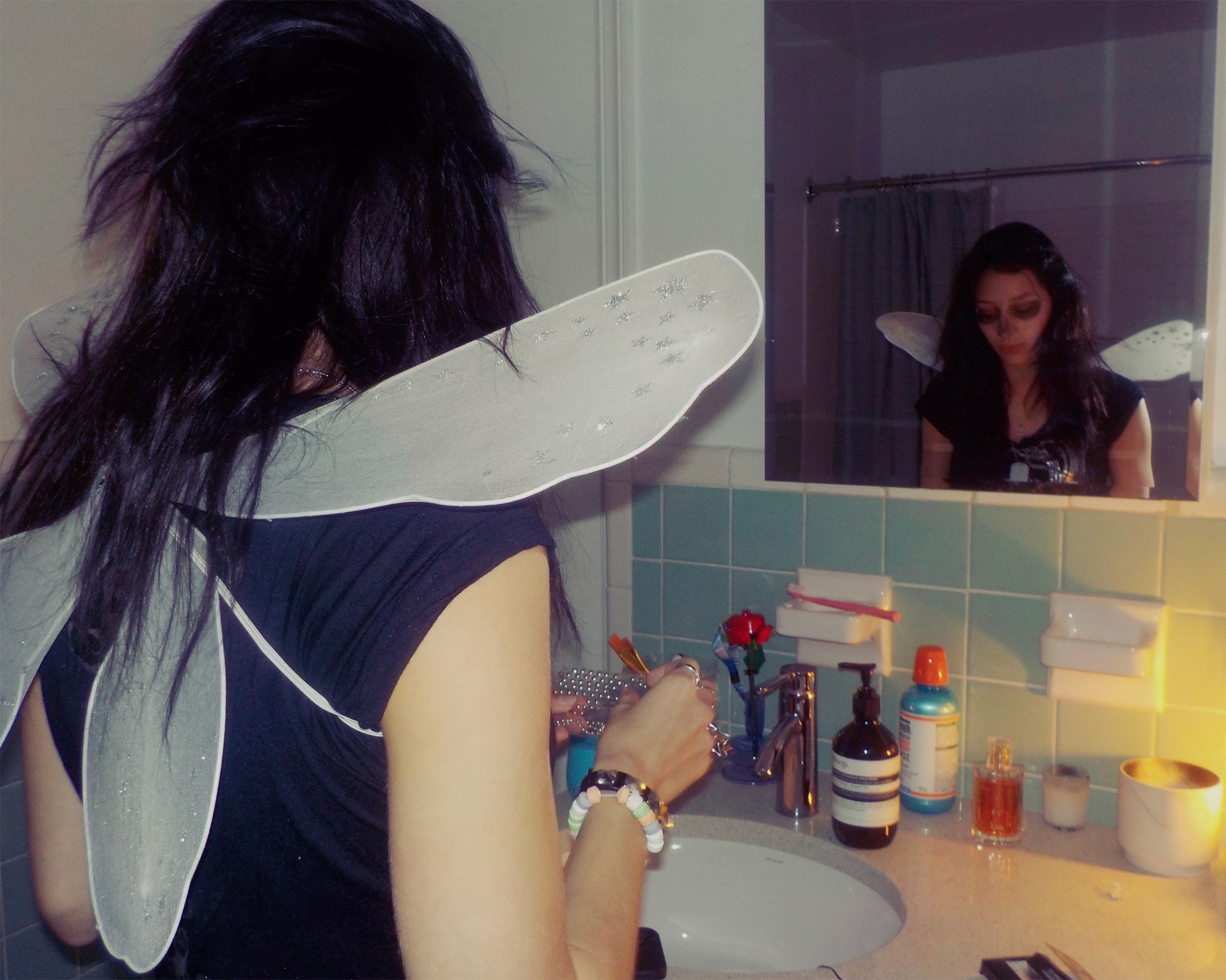 A young woman wears fairy wings and dark eye makeup, standing in front of a bathroom mirror.