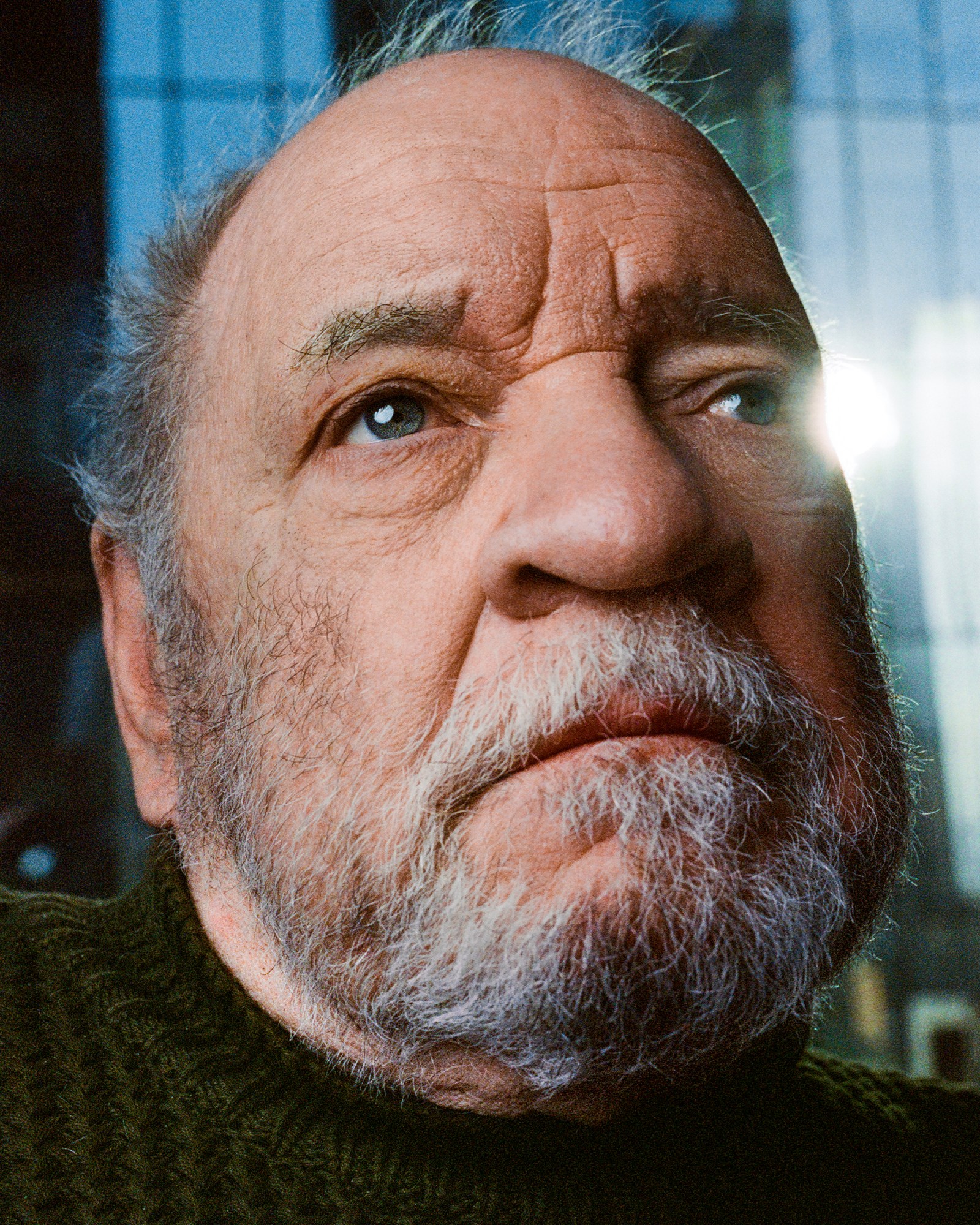 a close-up portrait of film director paul schrader, an elderly man with short grey hair and a beard. he's looking off camera as blue light streams through the window behind him