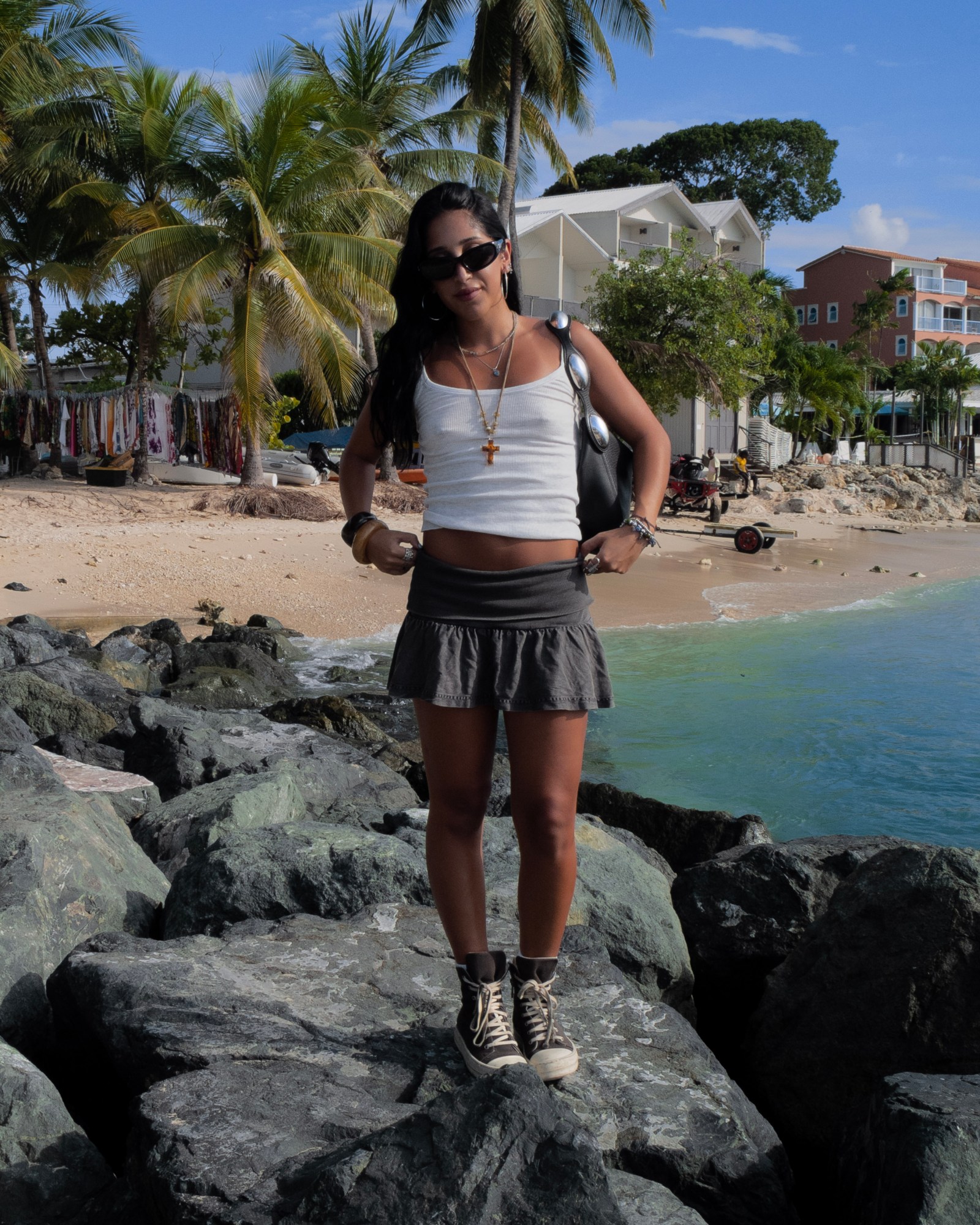 Woman in white tank top and pleated grey skirt stands on rocks next to the sea.