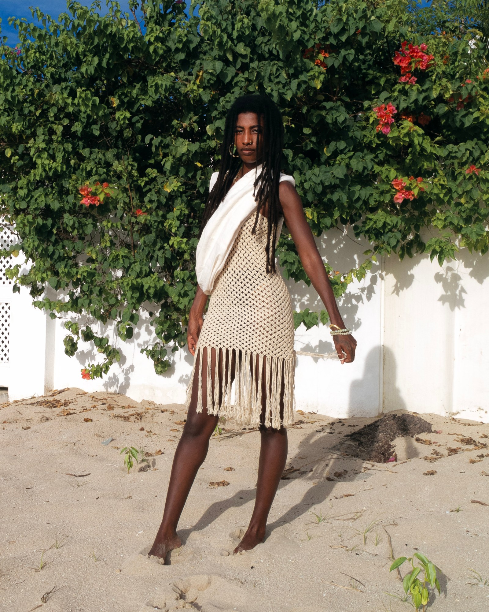 Woman in a beige knitted dress stands in front of a bush on the beach.