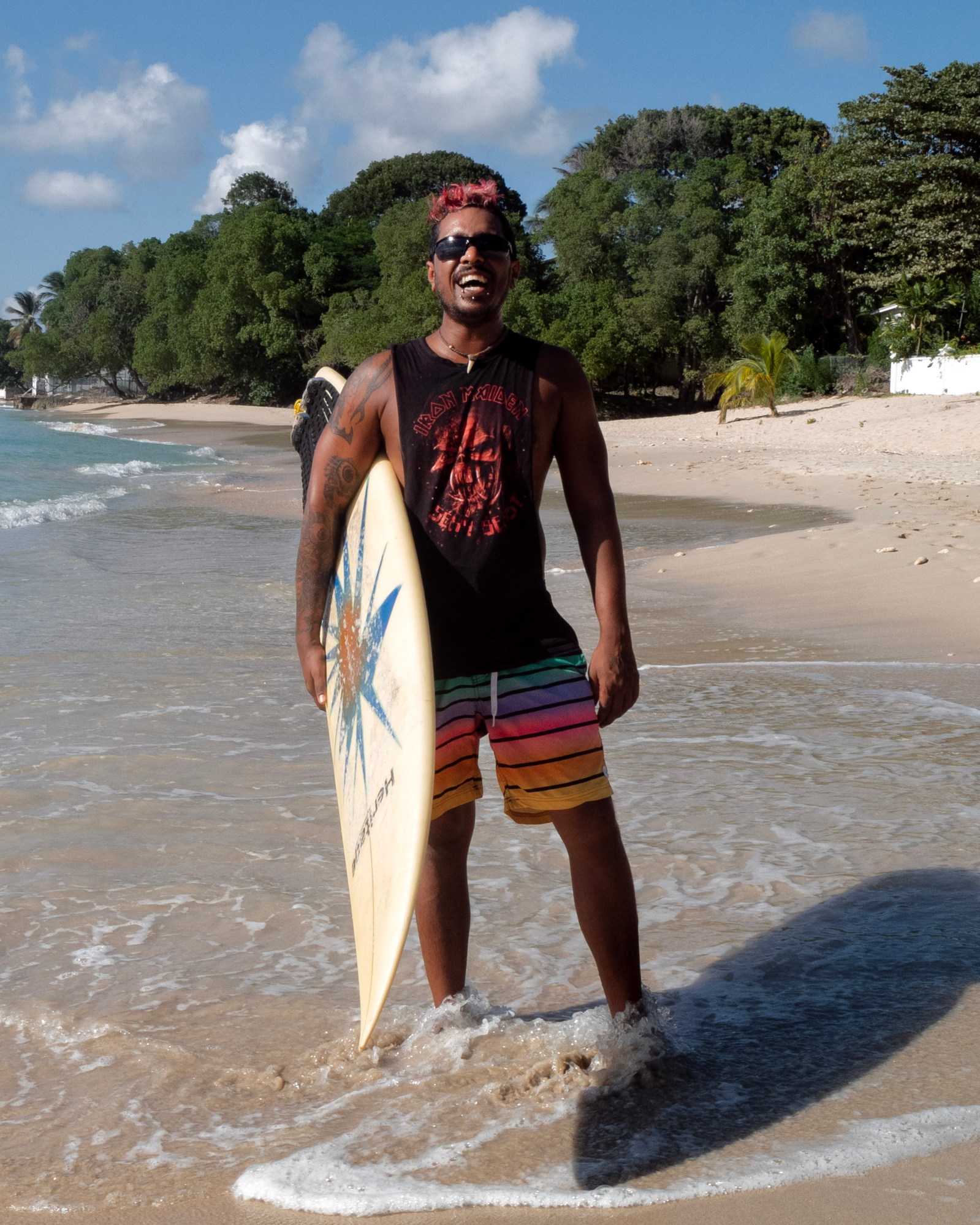 Man stands on the shore of the beach with surfboard wearing striped swimming shorts and black and red tank top.