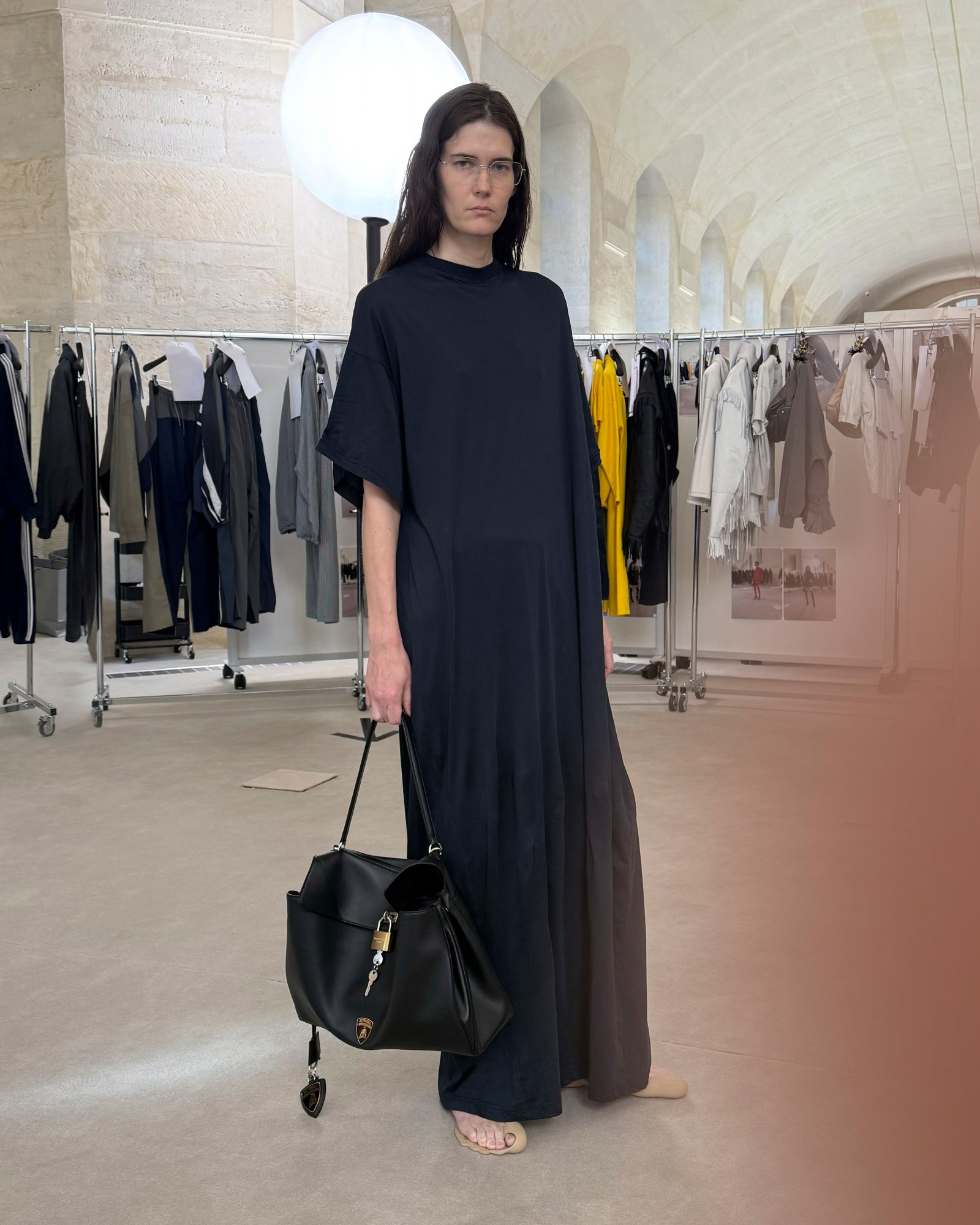 A model poses amongst clothing rails wearing a loose black dress and sand colored open-toed shoes