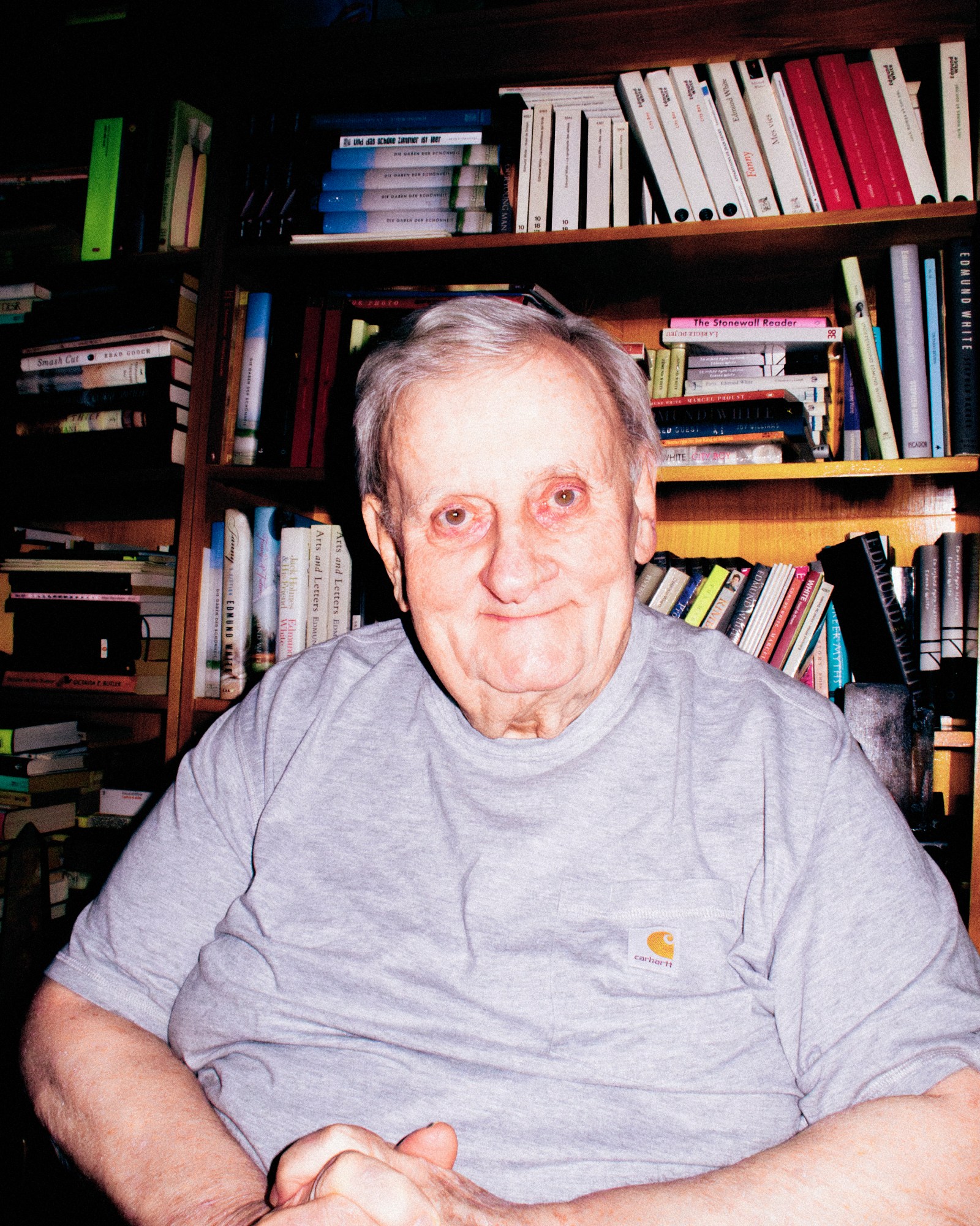 Edmund White author elderly gentleman wearing a grey t-shirt in his flat surrounded by a bookshelf