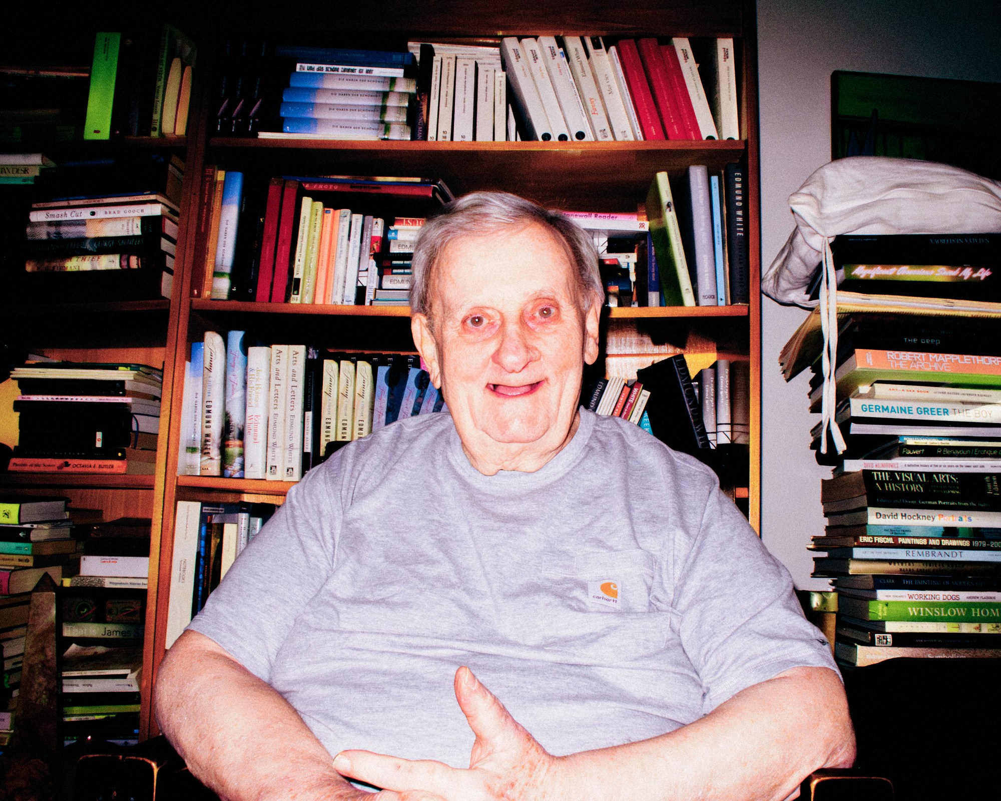 Edmund White author elderly gentleman wearing a grey t-shirt in his flat surrounded by a bookshelf