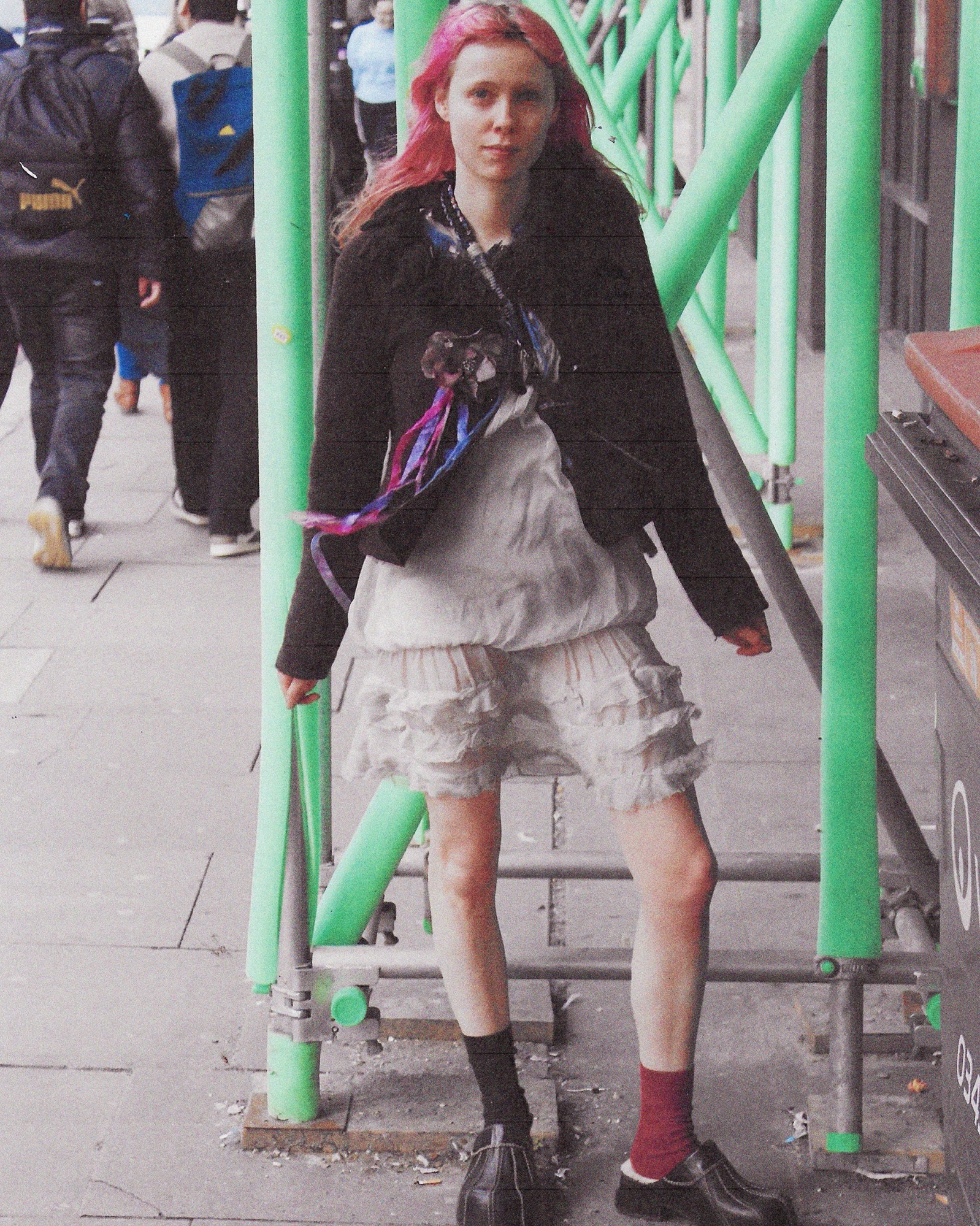 A full length photograph of a women with pink hear wearing a black jacket and grey dress in London.