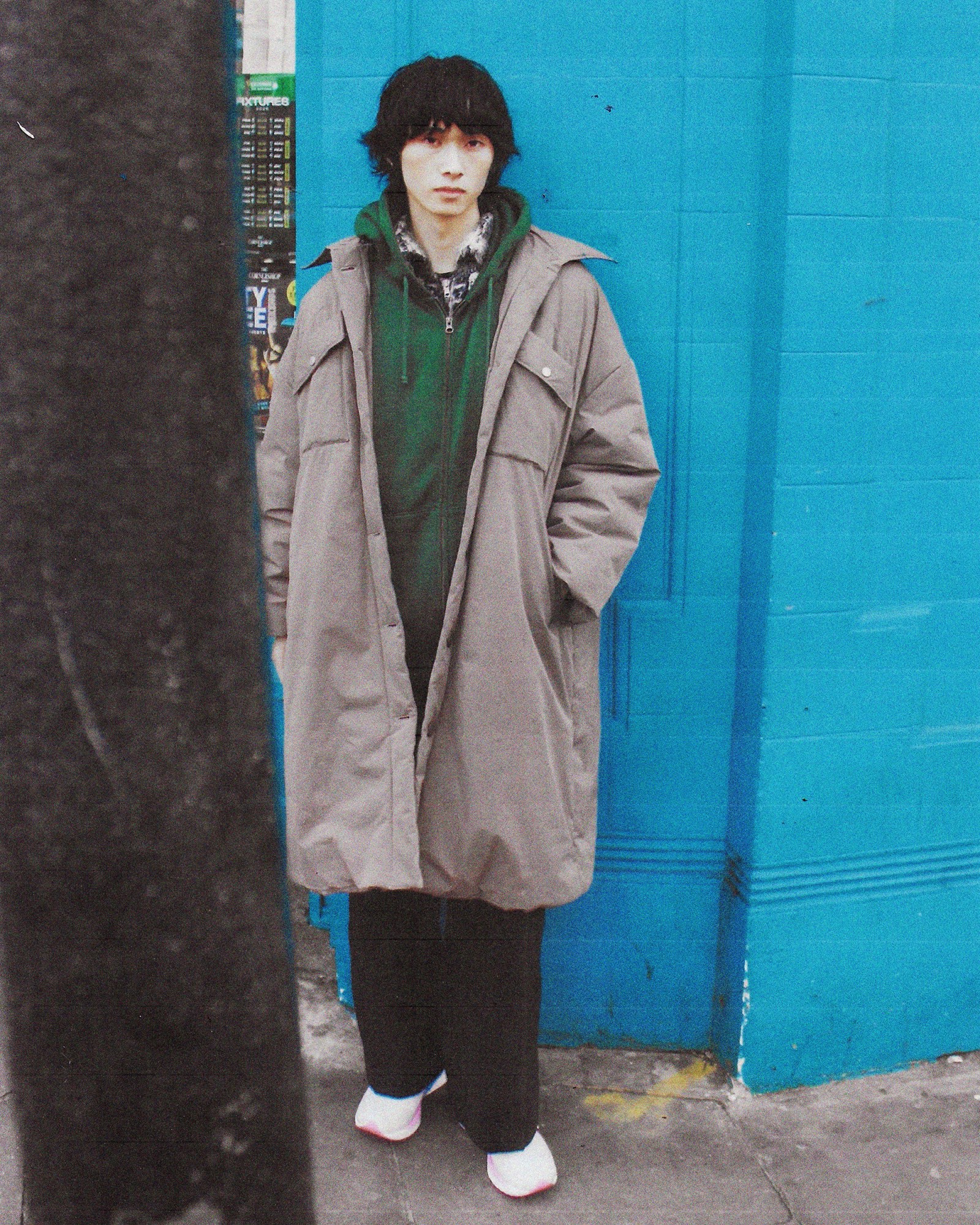 A full length photograph of a man wearing a grey jacket, green hoodie and jeans in London.