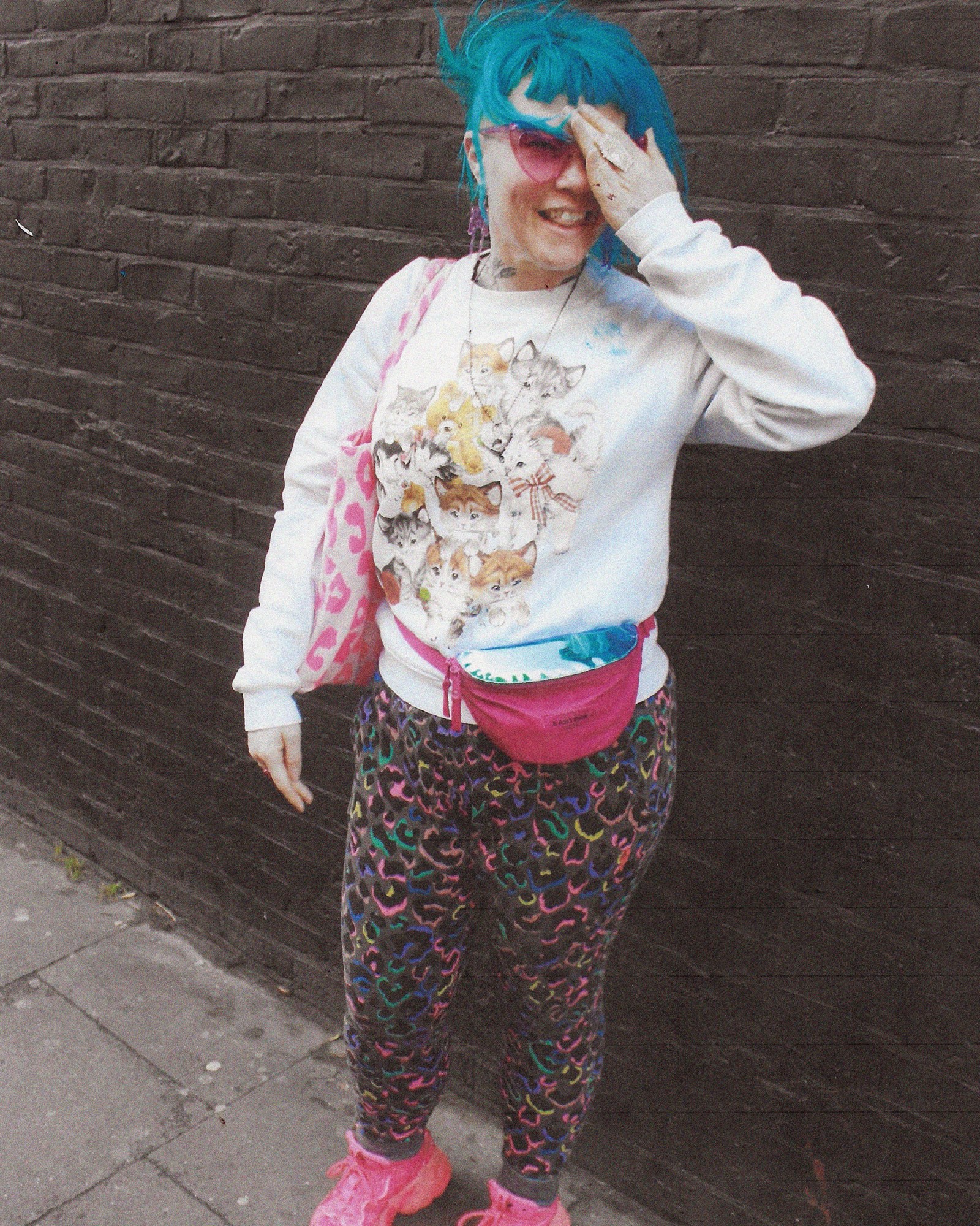 A full length photograph of a women with blue hair wearing a white jumper, multicolored leopard leggins and pink shoes.