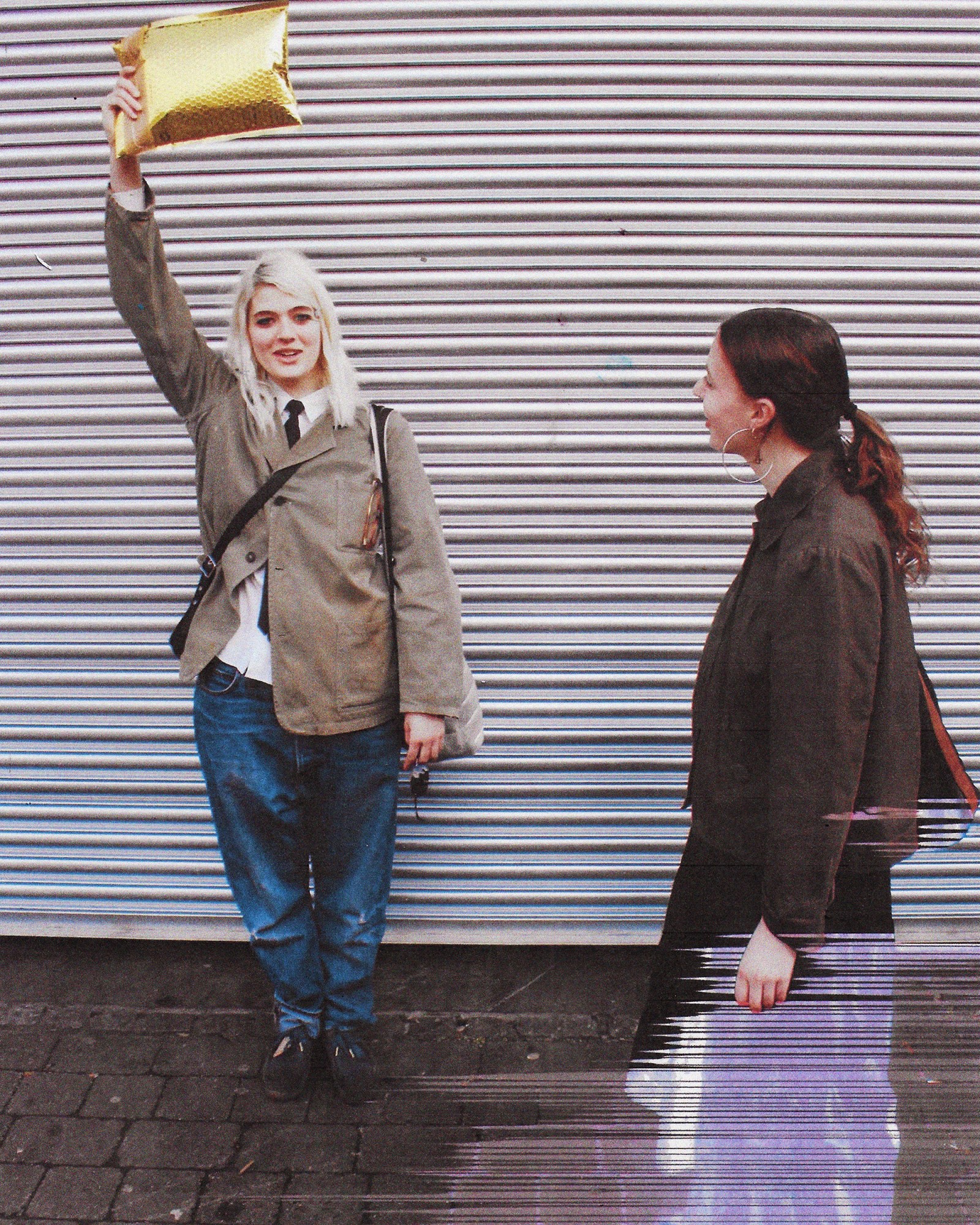 A full length photograph of two women in London, one holding a golden envelope above ehr head in a beige suit jacket and blue jeans, the other wearing a black jacket.