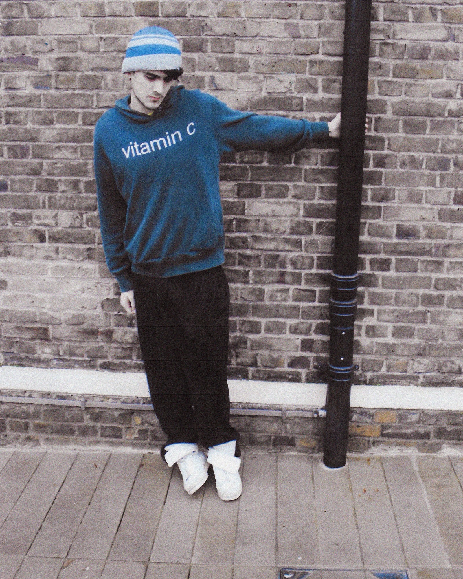 A full length photograph of a man wearing a beanie, a blue hoodie that says "vitamin C" and black jeans in London.