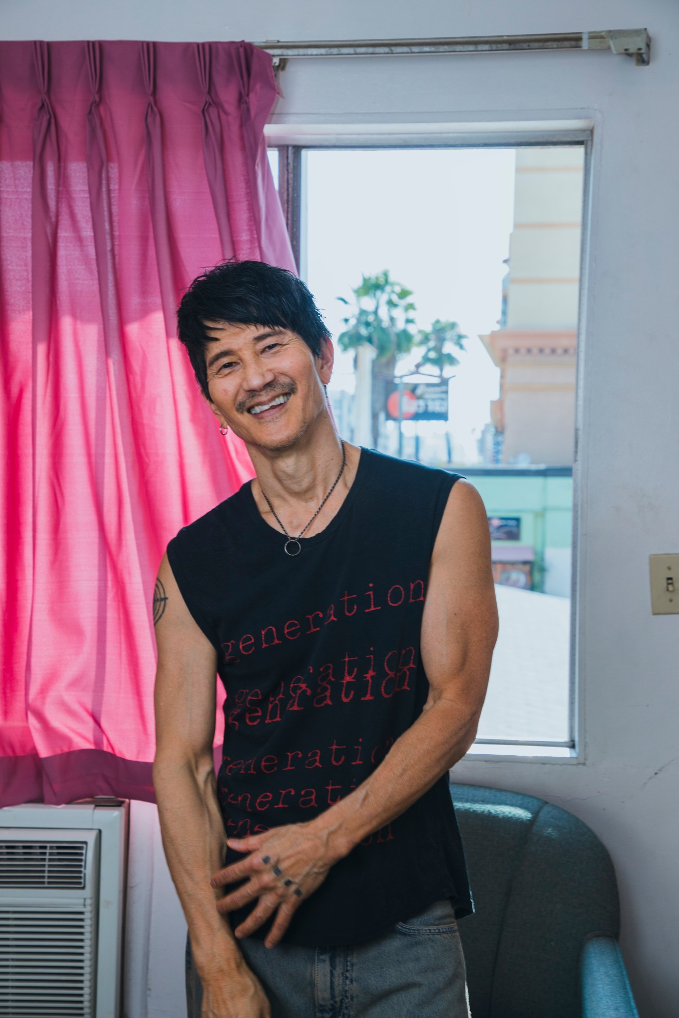 Gregg Araki by Kevin Amato for i-D magazine issue 375, fall/winter 2025. He's posing in front of a pink curtain wearing a black vest and smiling for the camera.