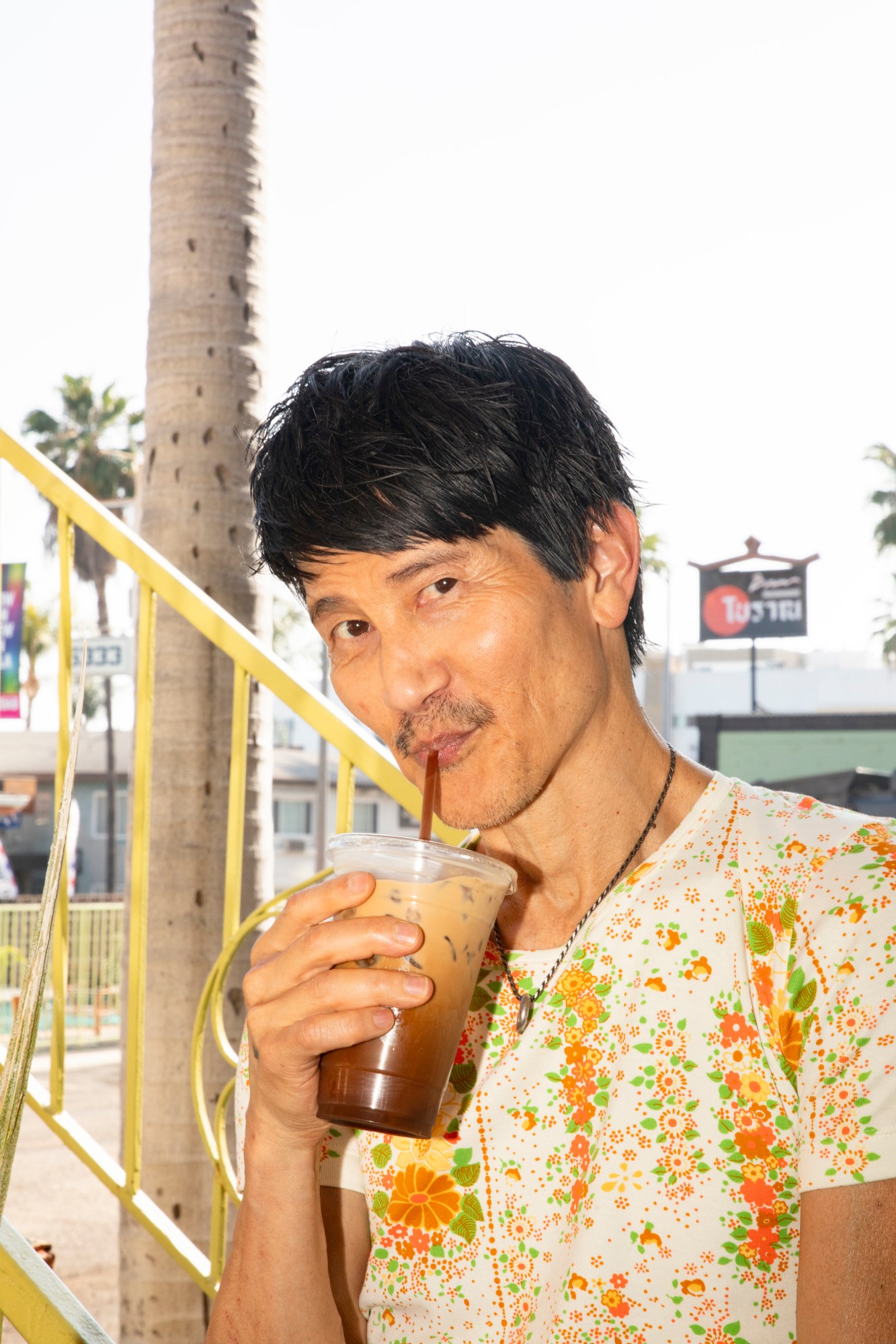 Gregg Araki by Kevin Amato for i-D magazine issue 375, fall/winter 2025. He's wearing a floral -shirt and is sipping an iced coffee.