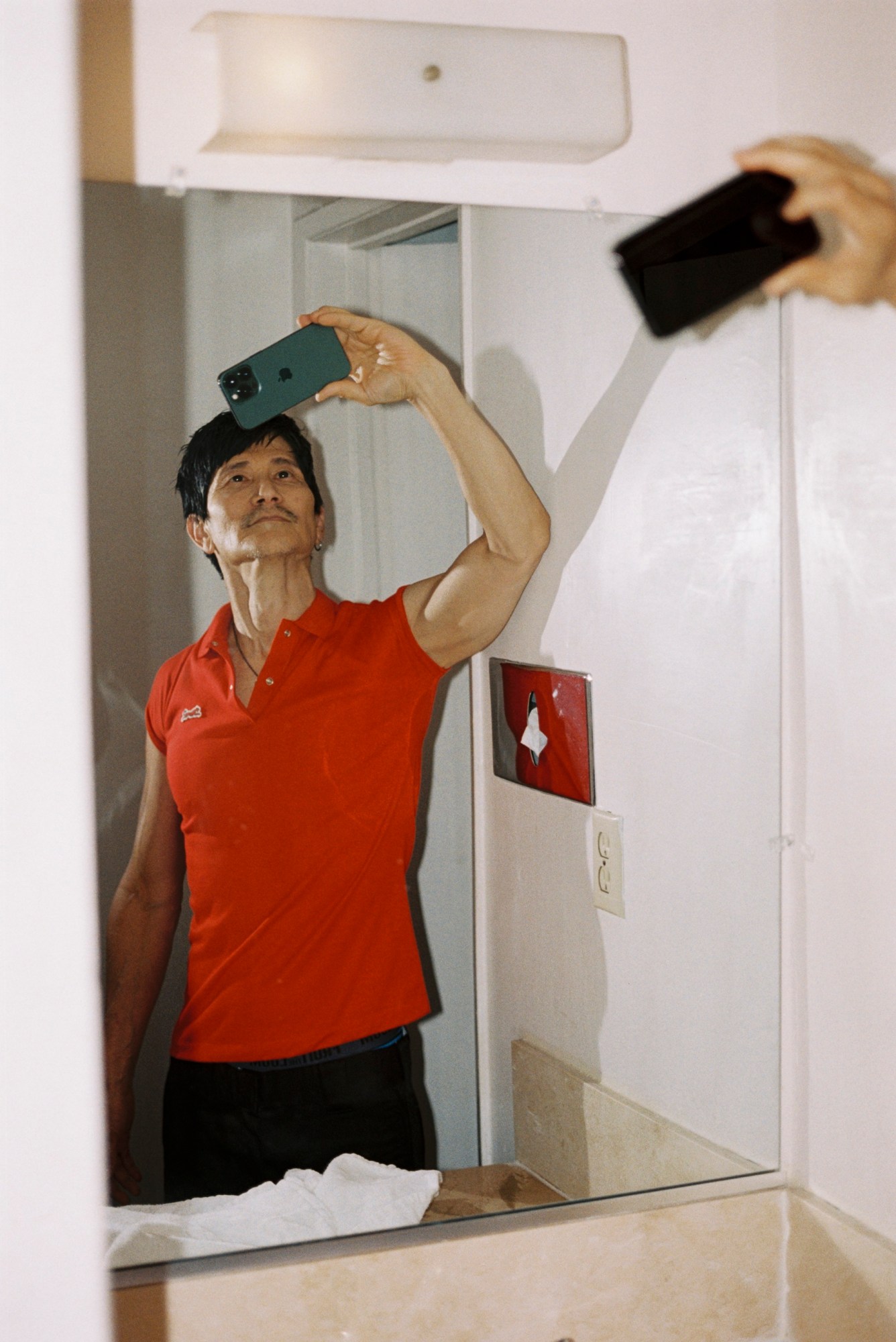 Gregg Araki by Kevin Amato for i-D magazine issue 375, fall/winter 2025. He's wearing a red polo shirt and is posing for a selfie.