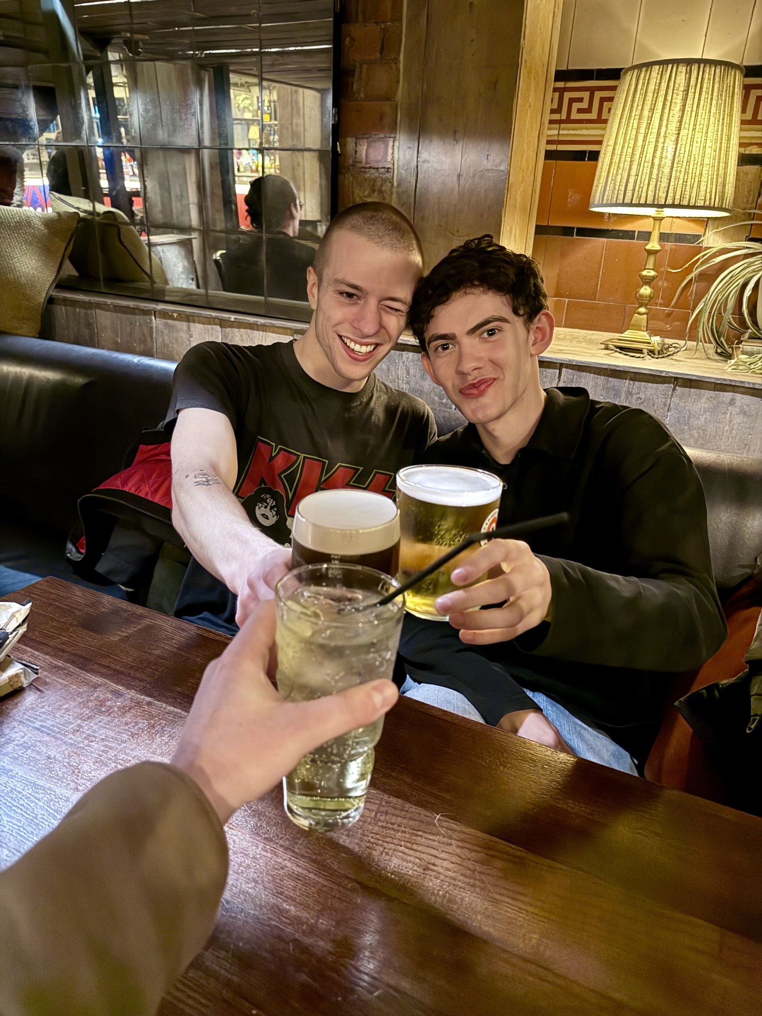 joe locke and ruaridh mollica at a pub in london. ruaridh wears a red jumper and has a shaved head. joe has curly dark hair and is wearing a dark jumper. they both appear in marvel shows, vision quest and agatha all along, as well as the play clarkston