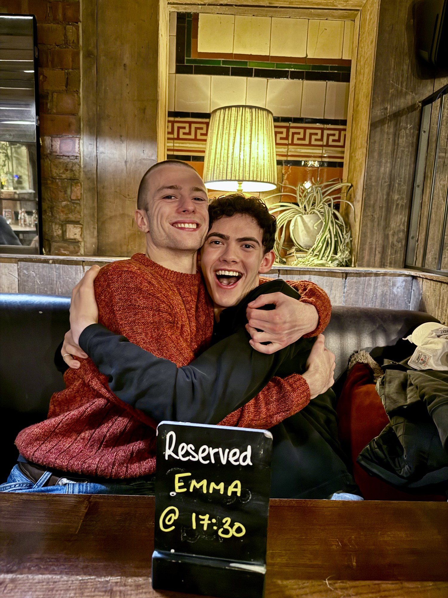 joe locke and ruaridh mollica kissing at a pub in london. ruaridh wears a red jumper and has a shaved head. joe has curly dark hair and is wearing a dark jumper. they both appear in marvel shows, vision quest and agatha all along, as well as the play clarkston